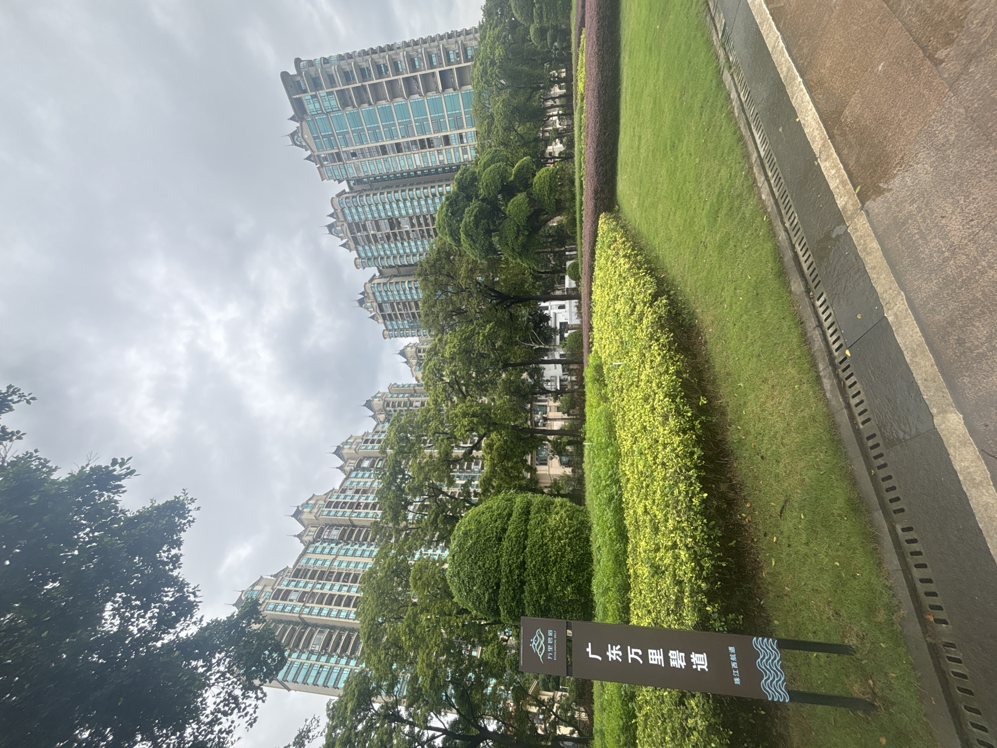 The image depicts a modern urban landscape in Nanhai District, China, under an overcast sky. In the background, several tall residential buildings with light-colored facades and blue-tinted windows dominate the skyline. These buildings feature ornate architectural details on their rooftops. In the foreground, a well-maintained park area includes lush green lawns, manicured hedges, and trees. A dark brown signpost with white text and a stylized wave logo is prominently displayed on the left side of the frame, indicating "Guangdong Wan Li Bi Dao" (Guangdong Thousand Mile Green Belt) and "Zhujiangxi Navigation" (Pearl River West Navigation). The park area is bordered by a paved walkway and a drainage system. The overall impression is one of urban development integrated with green spaces. No people are visible in the image.