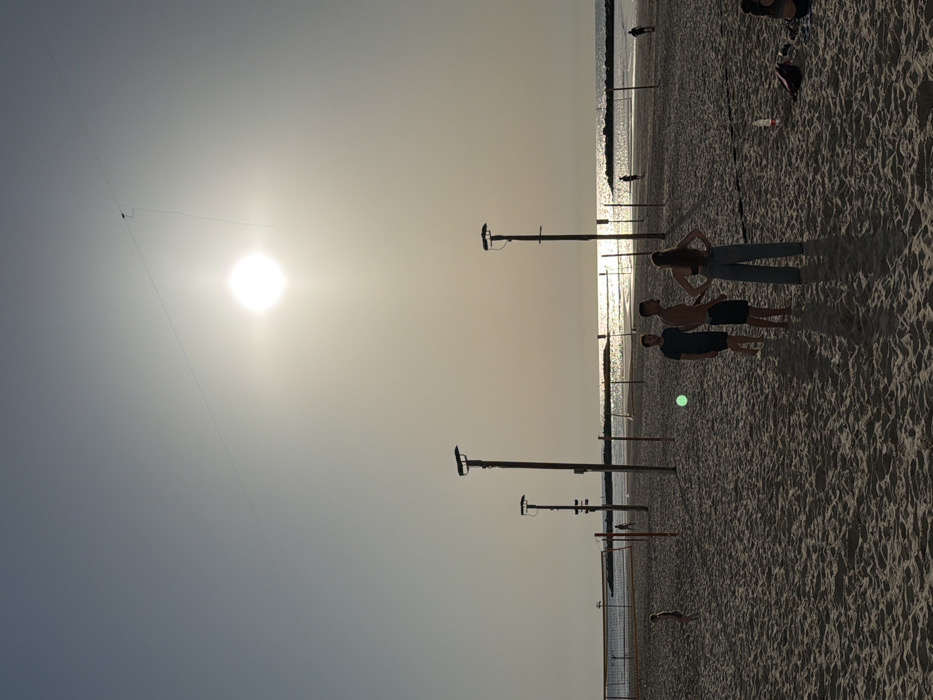 The image captures a late afternoon or early evening scene on a sandy beach in Tel-Aviv, Israel, with the low sun creating a bright glare over the water. The sky is a pale, hazy blue-gray, with the intensely bright, white sun positioned in the upper right quadrant. A thin line, identified as a kite string, extends diagonally from the upper right, near the sun, across the sky, with a small dark object (the kite) attached.
In the foreground and midground, a sandy beach occupies the lower portion of the frame. Multiple beach volleyball courts are established, marked by dark poles and nets that stretch across the sand. Several taller, dark poles, possibly acting as light standards, are also visible.
Three individuals stand in the mid-foreground. From left to right: one male wearing a dark t-shirt and shorts, another male shirtless and wearing dark shorts, and a female facing away from the camera, wearing a dark top and light-colored wide-leg pants, with her hands on her hips. A smaller figure, possibly a child, stands further back to the left, near a volleyball net. A green circular lens flare is present in the mid-left foreground. On the far right, partially cropped, another person is seated on the sand, with a red and white plastic bottle and a dark bag nearby.
In the background, the Mediterranean Sea reflects the bright sunlight, creating a shimmering effect. A stone breakwater or jetty is visible in the middle distance, extending into the water. Two additional indistinct figures are discernible on the wet sand near the water's edge, silhouetted against the bright reflection.