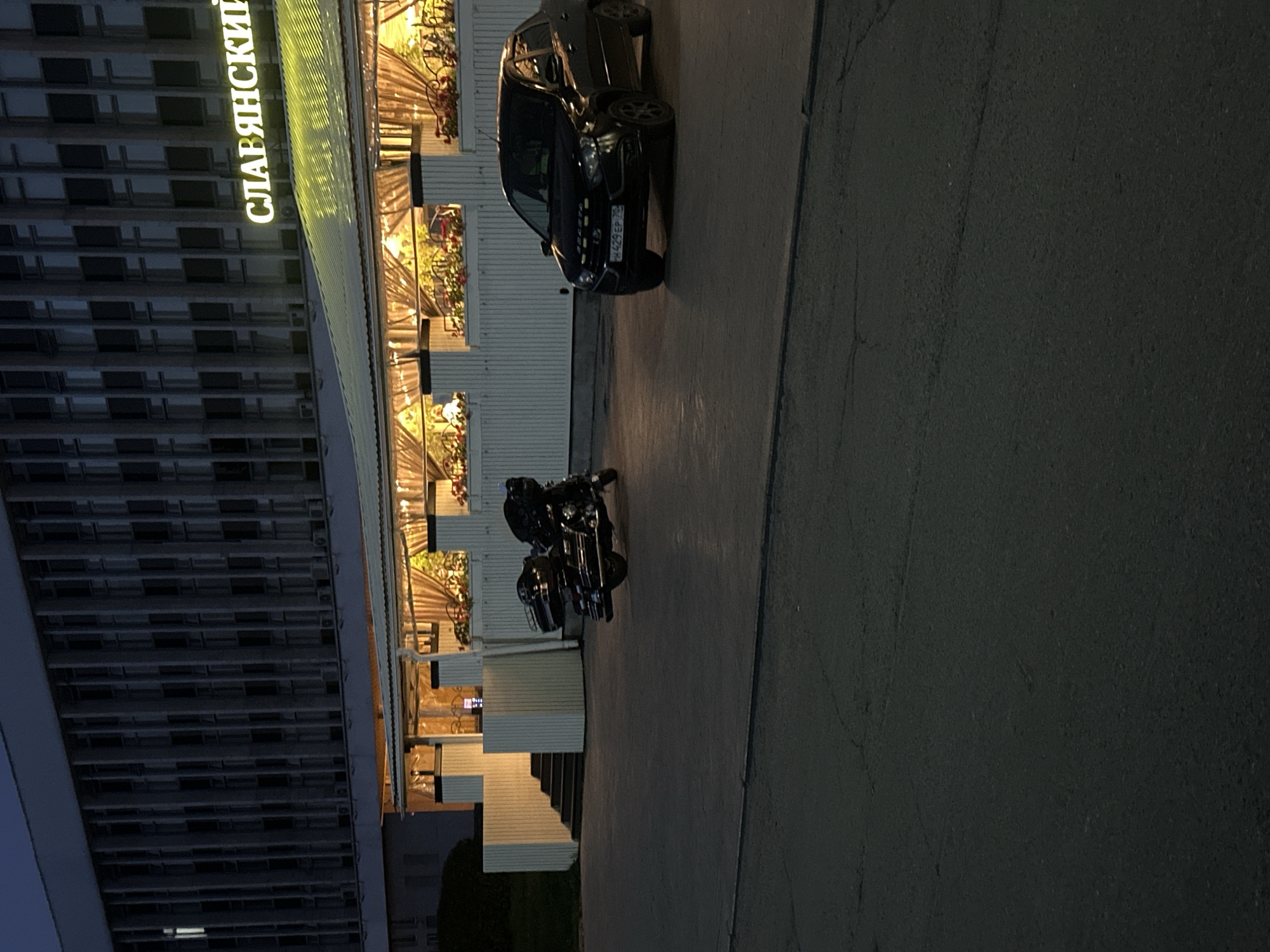 The image shows a nighttime exterior scene in Tomsk, Russia, featuring a building with a lit awning and a parking area. The building appears to be a restaurant or cafe, indicated by the illuminated interior visible through large windows and the sign that reads "СЛАВЯНСКИЙ" in glowing yellow letters. The awning is supported by white pillars, and decorative plants are visible along the railing.
In the foreground, a black sedan is parked on the asphalt. Its license plate, partially obscured, appears to be H429EP 70. To the left of the car, a black motorcycle, possibly a cruiser or touring model with saddlebags and a backrest, is parked. Both vehicles are stationary.
The environment is a paved outdoor area, likely a parking lot or sidewalk in front of the establishment. The lighting is dim, typical of evening or nighttime. The building facade is a muted gray with many vertical window sections. The ground is asphalt with some visible cracks.