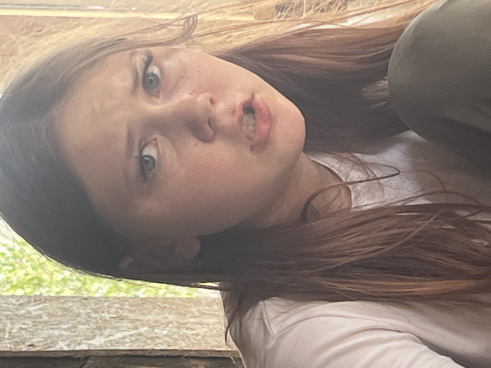 A young person with light brown hair and light-colored eyes is shown in a close-up, head-and-shoulders shot. They are wearing a white t-shirt and dark pants. The background consists of a wooden structure with vertical planks and blurred green foliage visible through a gap. The lighting is natural and appears to be from an outdoor source. The person's mouth is slightly open, revealing teeth, and their expression is ambiguous. The image quality is somewhat soft, possibly due to low light or motion blur. The location is described as Фоки, Russia.