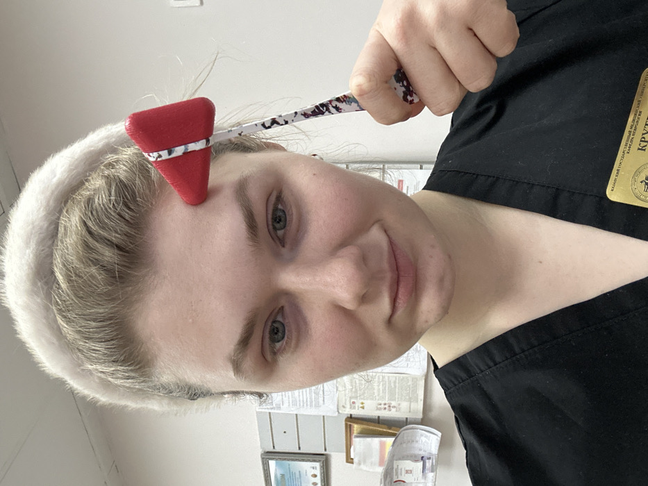 A fair-skinned woman with blonde hair and grey eyes holds a red reflex hammer to her forehead. She wears a white headband and black scrubs with a badge. The background shows a cluttered wall with papers, a picture frame, and rolled-up paper. The image is slightly rotated to the left.
