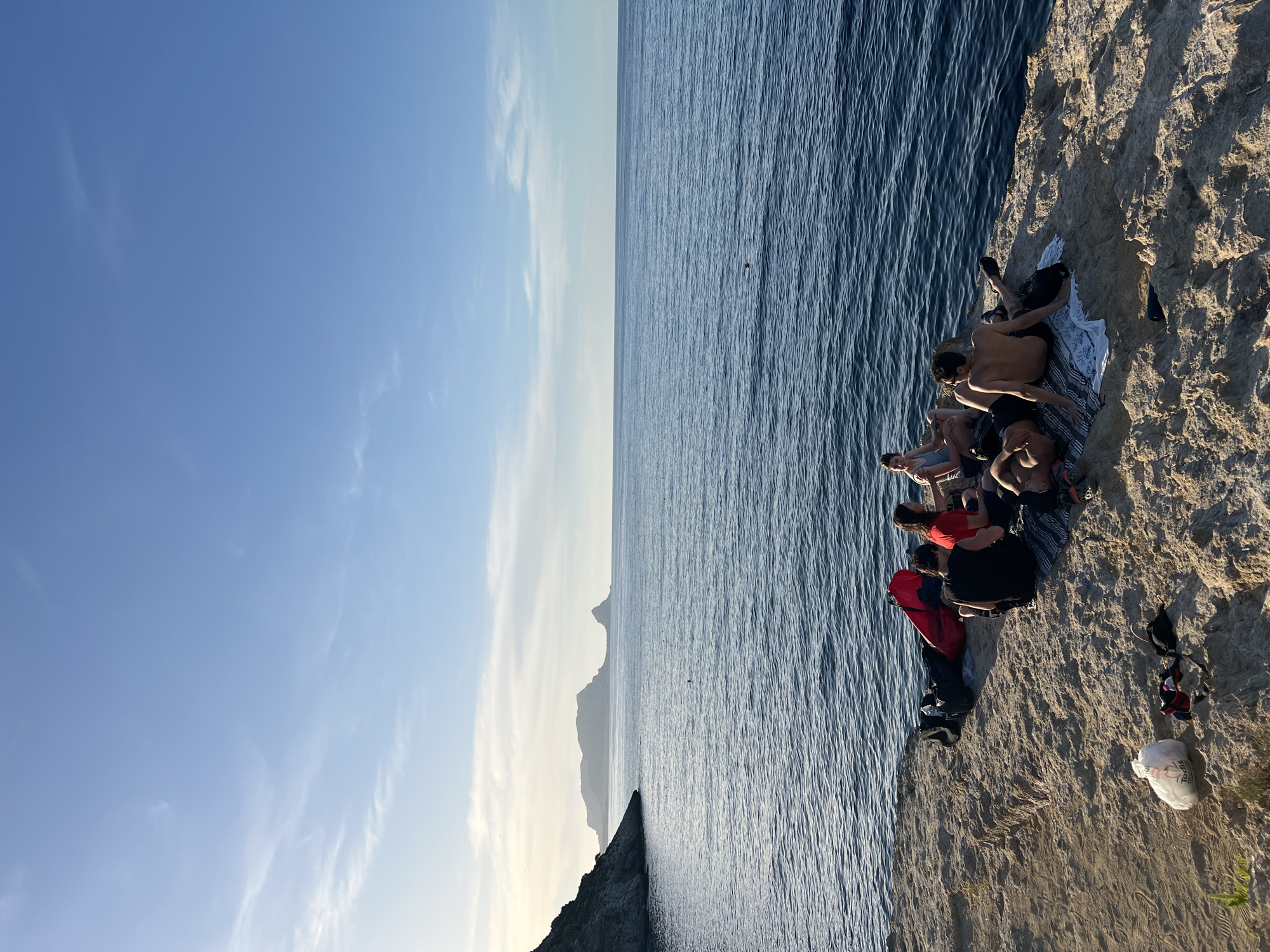 The image depicts a coastal scene in Sperone, Italy, at what appears to be late afternoon or early evening, characterized by warm, low-angle sunlight. The sky is predominantly clear blue, with scattered, wispy white clouds concentrated on the left horizon, which is lighter in tone. The sea, a calm deep blue, shows gentle ripples across its surface. In the mid-ground, a dark, indistinct object is visible in the water.
Across the distant horizon, a series of dark, silhouetted landmasses or mountains rise from the sea, with a more pronounced peak on the right. The foreground features a light-colored, textured rocky outcrop or cliff face, where a group of approximately seven individuals are gathered. The rock surface is uneven, showing shadows consistent with the sun's low position. Sparse green vegetation is visible on the far left edge of the outcrop.
The individuals are arranged in small clusters on blankets or towels spread on the rock. From left to right:
- A red duffel bag and a dark backpack are placed on the rock.
- A person in a black t-shirt sits with their back to the viewer, facing the sea.
- To their right, a person in a red t-shirt sits, also facing the sea.
- Further right, two individuals are relaxing on a striped blanket. One person (bare-chested) lies on their stomach, facing away from the camera. Another person (bare-chested male) sits upright with their back to the camera, wearing dark shorts and shoes.
- On the far right, two more individuals are seated. One person in a light top sits with their back to the camera, and another person with dark hair sits facing the sea.
- A white plastic bag, bearing the text "Todis," rests on the ground in the far left foreground, alongside scattered items of clothing and footwear. A dark, small object (possibly a speaker) is visible on the rock near the central group.
The individuals appear to be relaxing and conversing in a casual manner, enjoying the coastal environment.