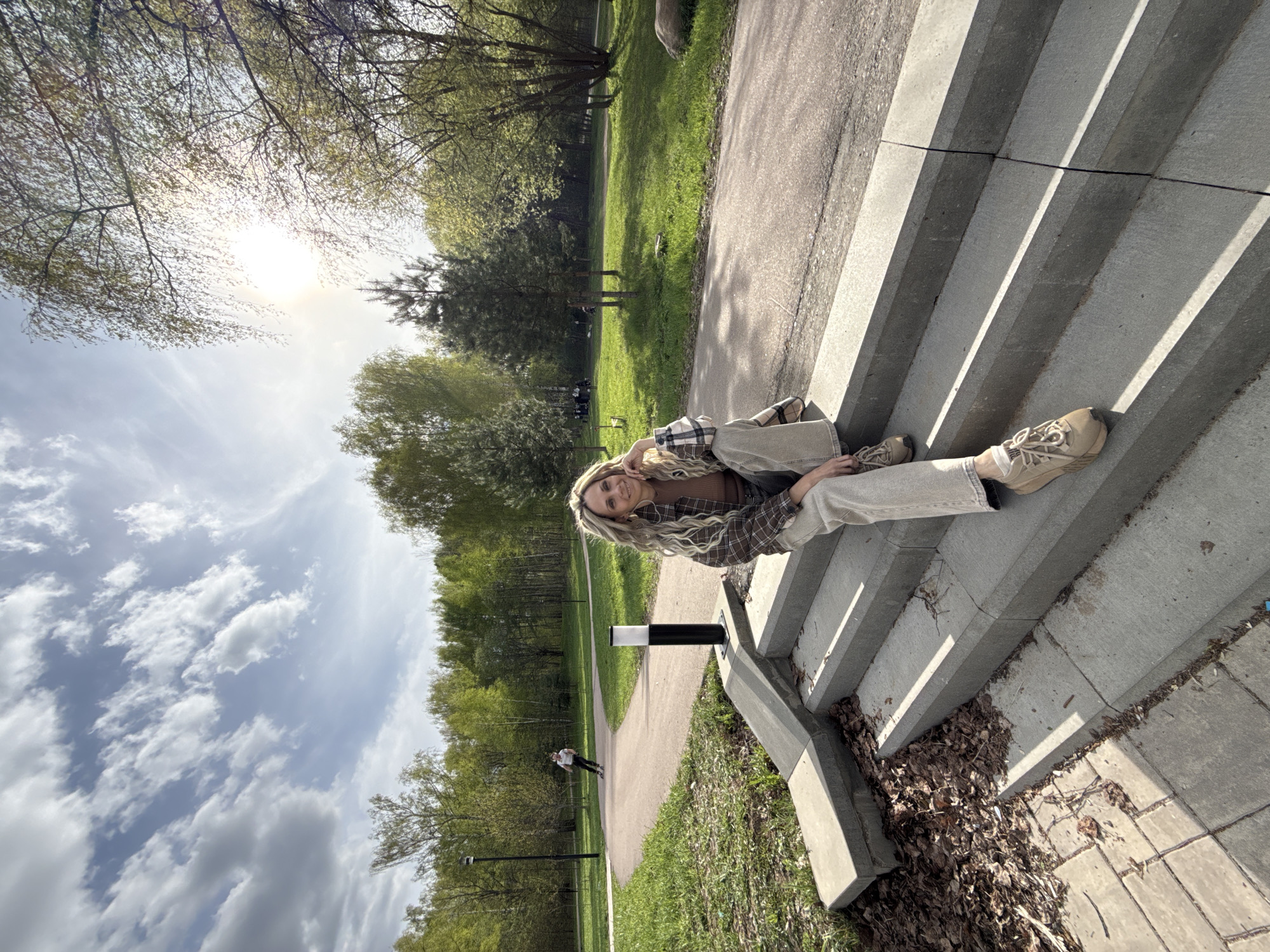 A young woman with long, blonde, curly hair is sitting on a set of concrete stairs in Rusanovo, Russia. She is wearing a brown shirt, a plaid jacket, and light-colored pants with tan sneakers. The stairs are bordered by grass and a walkway leading into a park with trees. The sky is partly cloudy with the sun shining brightly.