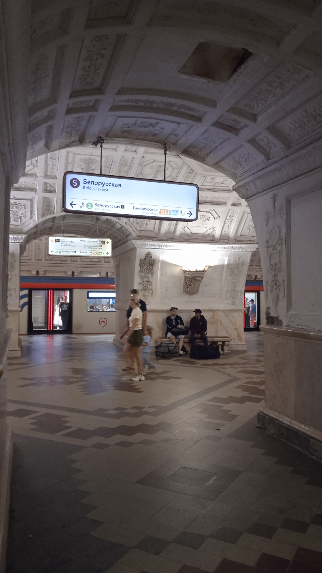 The image depicts a view within a Moscow Metro station, likely Belorusskaya station, as indicated by the prominent signage. The station features ornate architectural details, including arched ceilings with decorative plasterwork and marble-clad pillars.
In the foreground, a tiled floor with a geometric pattern is visible. Several individuals are present: a family consisting of two adults and a child is walking across the platform. Two men are seated on a bench in the middle ground, one wearing a dark cap and the other a baseball cap, with luggage beside them.
A train with illuminated windows is stopped at the platform on the left side of the image. The train is light-colored with blue and red stripes. Another train is partially visible on the right platform.
Directional signs, written in Russian, are prominently displayed, indicating "Belorusskaya" with line numbers and arrows. The lighting appears to be a mix of ambient station lights and the illumination from the trains. The overall impression is a typical scene in a busy Moscow Metro station.