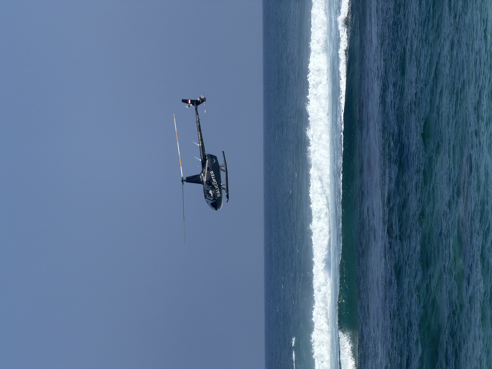 A black helicopter is flying over the ocean. The helicopter has the word "BALICOPTER" written on its side. The ocean is blue with white waves. The sky is clear and blue. The image appears to be taken from a distance, as the helicopter is small in the frame. The helicopter is in focus, while the ocean and sky are slightly blurred. The lighting suggests it is daytime, possibly midday, given the bright sky and the way the light reflects off the water. The scene is serene and suggests a recreational or transport activity over the water.