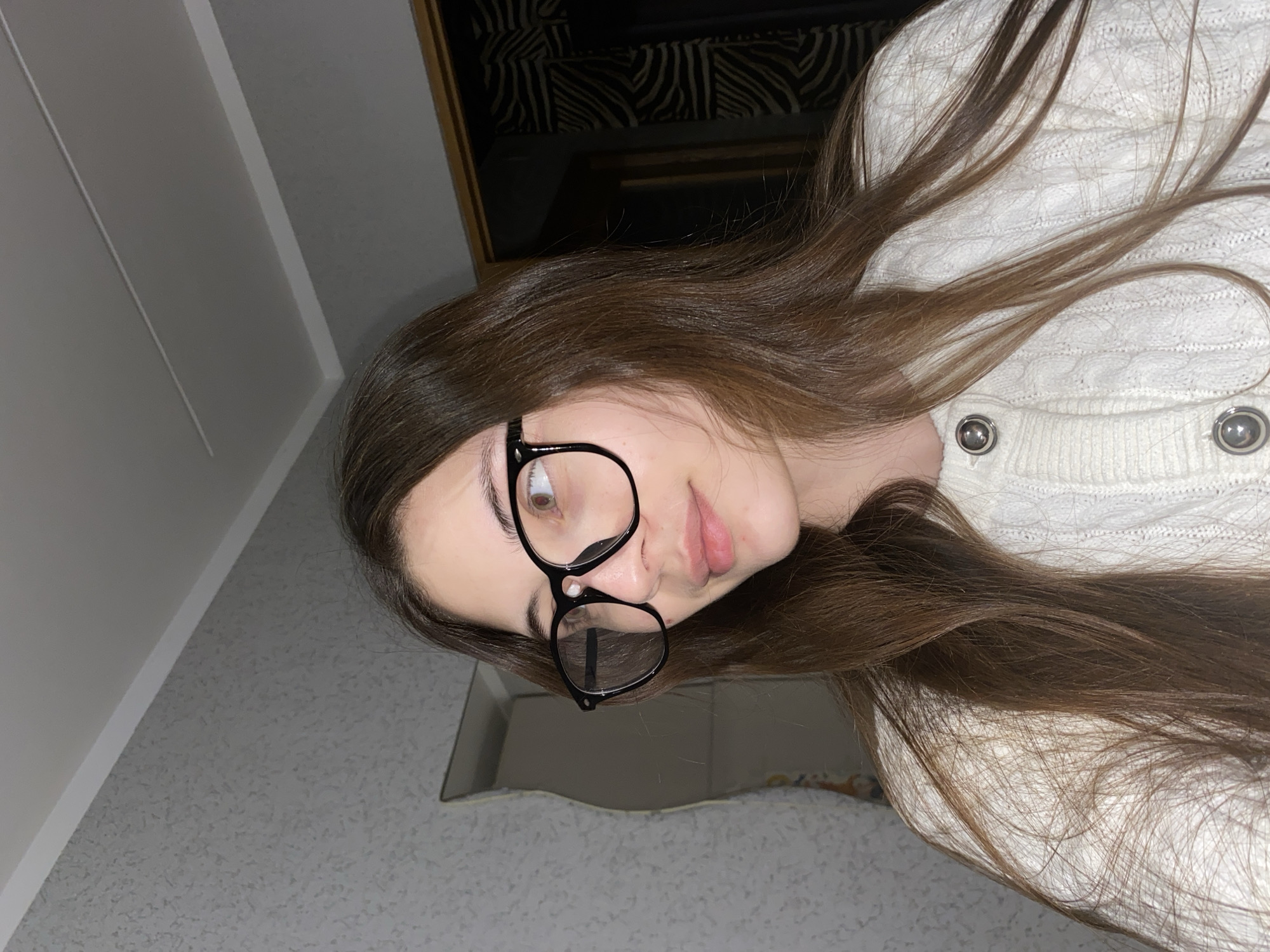 A close-up, rotated image captures a young woman from Votkinsk, Russia. She has long, straight, dark brown hair that cascades over her shoulders and a white cable-knit cardigan. The cardigan features a textured pattern and two prominent silver buttons are visible on the front. She wears black-rimmed, round spectacles. Her face is turned slightly, and her lips are softly pursed, giving her expression a thoughtful or slightly playful quality. Her gaze is directed upwards and to her right.
The background suggests an indoor setting. To the left and below her, a light gray wall with a subtle textured pattern is visible. Above and to her right, a darker area features a distinct black and white zebra stripe pattern, possibly wallpaper or fabric, framed by a wooden border. The overall lighting is soft, suggesting an indoor environment. No text is visible in the image.