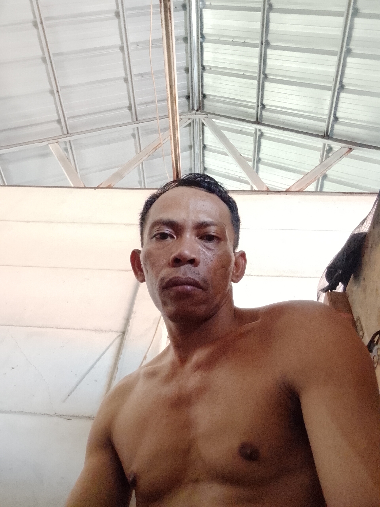 A shirtless man is looking directly at the camera. He has short, dark hair and his skin tone suggests he is Indonesian. The setting appears to be a room with a corrugated metal roof and walls, suggesting a simple dwelling. Based on the query of Bukit Ketok, Indonesia, the scene likely takes place there. There are no apparent actions or interactions beyond the man looking at the camera.