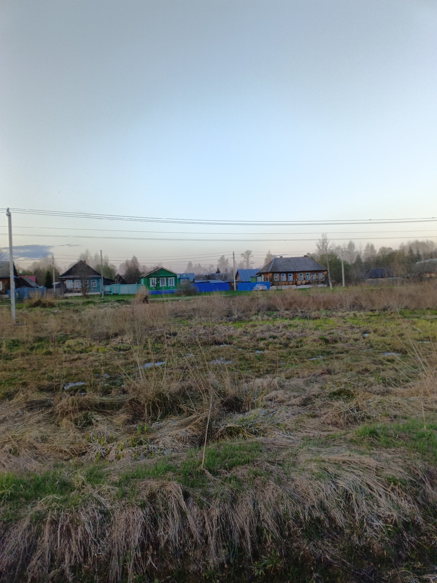 The image depicts a rural scene in or near Красный Маяк, Russia. In the foreground is a field of mixed brown and green grasses. Patches of bare earth or possibly residual snow are visible amongst the grasses. Beyond the field, a row of houses with varying architectural styles and colors, ranging from green to brown, can be seen. Some houses are fenced in. Power lines run horizontally across the upper portion of the scene. A line of trees is in the background. The sky is overcast and a light blue-gray color. The scene seems quiet and undisturbed.