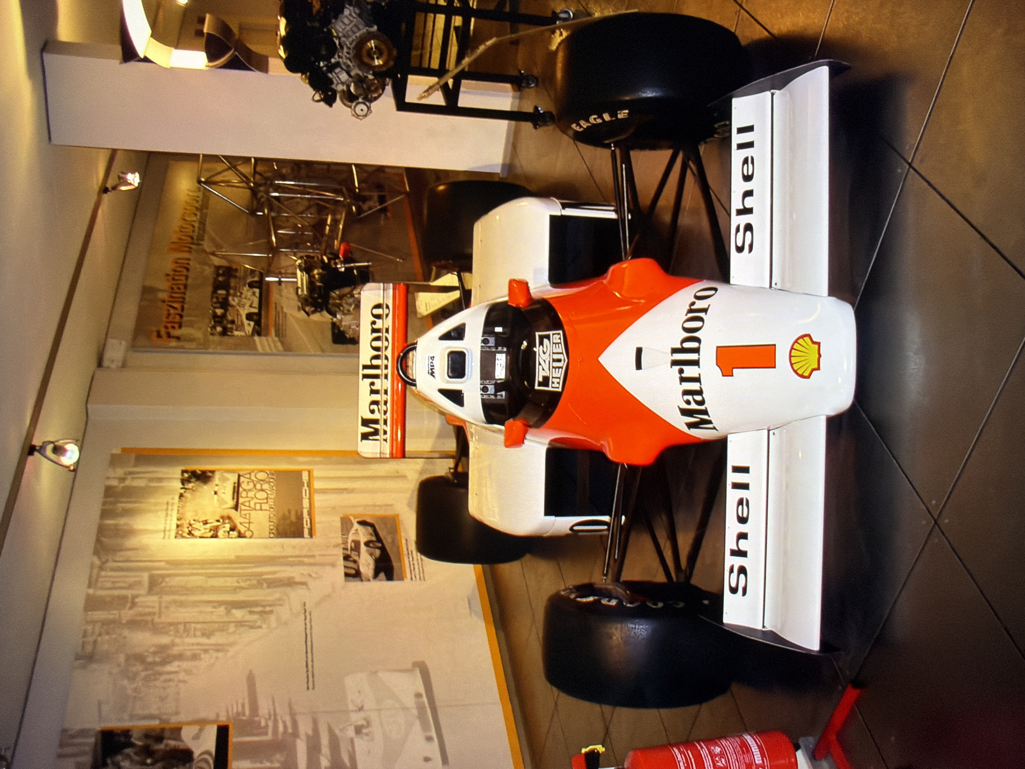 The image shows a close-up, eye-level shot of a Formula 1 racing car, predominantly white and red, parked indoors. The car features prominent "Marlboro" branding on its front wing and body, along with the number "1" and a Shell logo. The rear wing has "Shell" printed on it. The car appears to be on display, with its tires visible. In the background, there are framed posters on the wall, one of which appears to be an advertisement for "Faszination Motosport." To the right, a portion of an engine on a stand is visible. The lighting suggests an indoor exhibition or museum setting. The floor is tiled in a dark, reflective material. The image's metadata does not specify a location in Kyiv, Ukraine, or any other city.