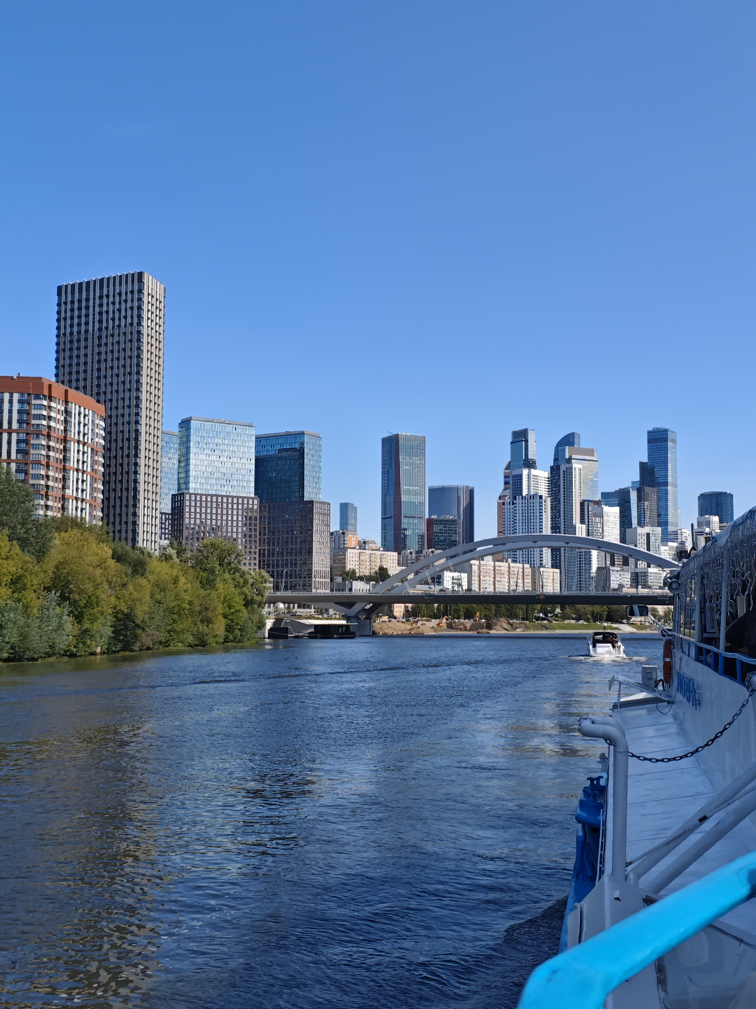 This is a vertical, eye-level, medium shot photograph taken from a boat moving down a wide, blue river in Moscow, Russia. The river dominates the lower two-thirds of the frame, with gentle ripples reflecting the clear, bright blue sky. On the left bank, a dense line of lush green trees and foliage separates the water from a collection of modern, high-rise buildings. These buildings vary in height and architectural style, with some featuring glass facades and others with more traditional window patterns.
A distinctive arched bridge spans the river in the mid-ground, connecting the two banks. Beyond the bridge, a cluster of very tall skyscrapers, characteristic of Moscow's Moscow City business district, rises against the horizon. These buildings are a mix of contemporary designs, with some clad in glass and others in lighter materials.
In the foreground, on the right side of the image, the white and blue side of a large passenger boat is visible. A blue railing runs along its edge, and a metal chain hangs from it. Further down the river, a smaller white motorboat is seen moving away from the camera, leaving a white wake. The overall scene depicts a sunny day with good visibility, showcasing the juxtaposition of nature and modern urban development along the Moscow River.