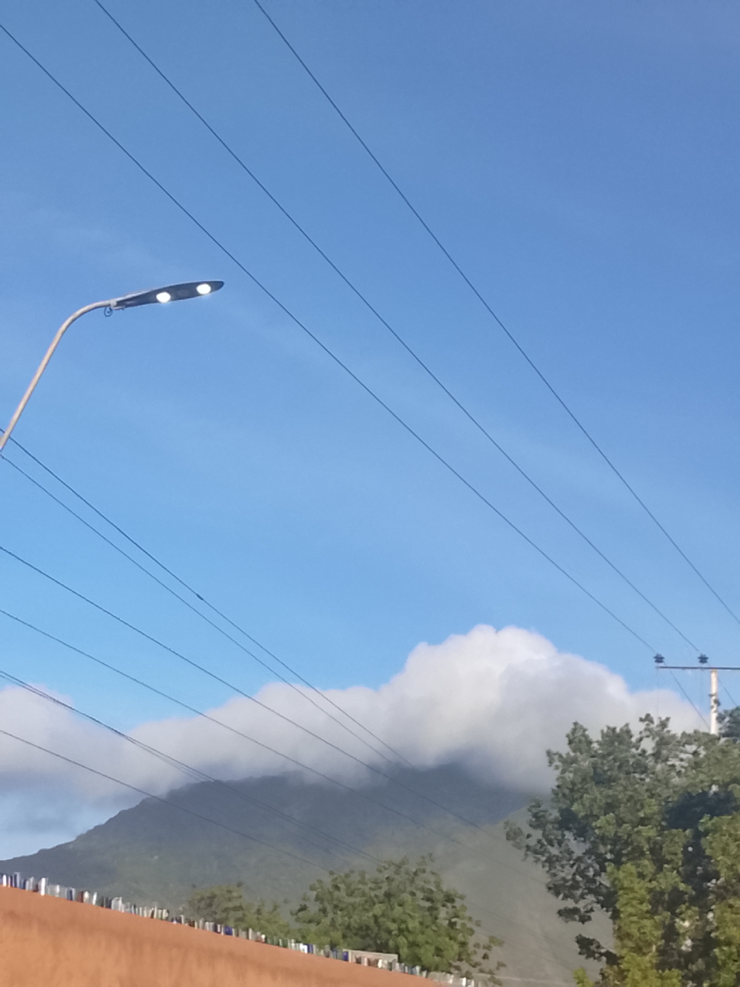 A daytime exterior shot in Altagracia, Venezuela, shows a street light with two illuminated bulbs against a clear, blue sky crossed by multiple power lines. Below, a partially cloud-covered mountain is visible. In the foreground, there is a low structure, possibly a fence or building, with sporadic greenery and trees to the right. The view suggests an urban fringe area where the town meets the natural landscape.