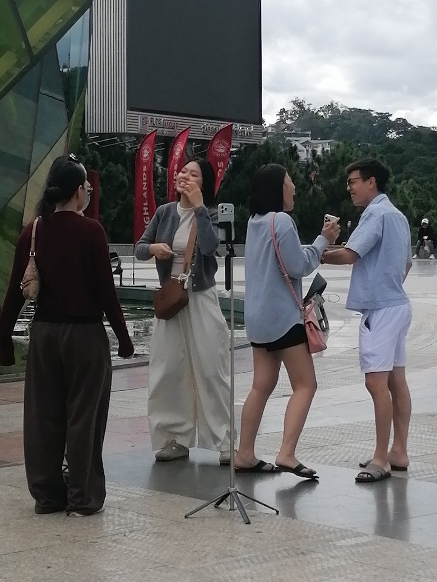 A group of four individuals are standing in an outdoor, paved area with a large building and a hillside with trees and structures in the background. The scene appears to be in Da Lat, Vietnam.
On the far left, a person with dark hair tied back in a bun, wearing a dark maroon long-sleeved shirt and loose-fitting dark brown pants, stands facing away from the camera. They have a beige purse with a strap over their left shoulder and a white mask is visible below their chin.
To their right, a young woman in a grey cardigan over a white top and a long, flowing white skirt is facing left and eating something. She has a small, brown heart-shaped purse hanging from her left shoulder. Her mouth is open, and her hand is near her face.
In the center-right, a young woman wearing a light blue button-down shirt, black shorts, and a pink crossbody bag is facing the person to her right. She is holding up a smartphone, possibly taking a photo or video. She has on black sandals.
To her right, a young man in a light blue short-sleeved button-down shirt and white shorts is facing the woman in the blue shirt. He is holding up a smartphone and appears to be looking at it or interacting with the woman. He is wearing glasses and grey sandals. A black tripod is visible between the two middle individuals, with a smartphone mounted on it.
The environment features a large building with a dark screen or display panel and architectural elements on the left. Red flags with text are visible in the mid-ground. Beyond the paved area, there is a lush green hillside with trees and some buildings. The sky is overcast. The ground is paved with light-colored tiles or stones, and there are some reflective surfaces.