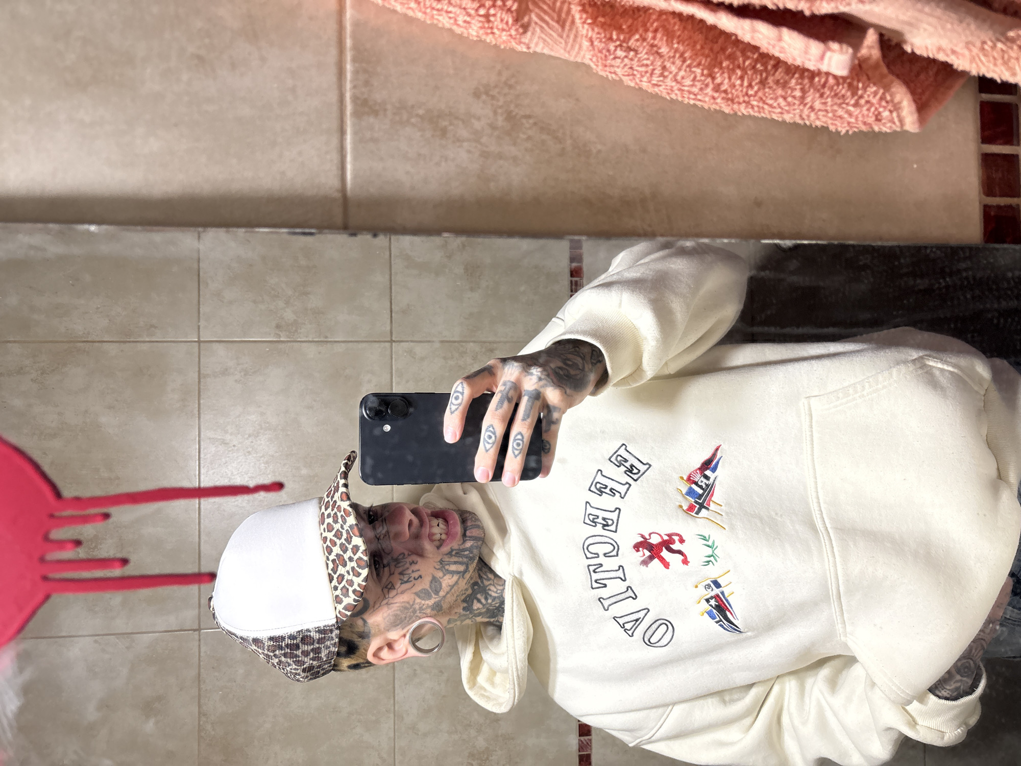 A tattooed individual is taking a selfie in a bathroom. The person is wearing a white hoodie with the word "OVERCITAO" and flags embroidered on it, a leopard print brim hat, and has gauges in their earlobes. Their face and hands are heavily tattooed. They are holding a black smartphone. The background features tan tiles, a pink towel, and a red splatter-like design on the mirror. The location is likely a bathroom in an unspecified building in Buenos Aires, Argentina.