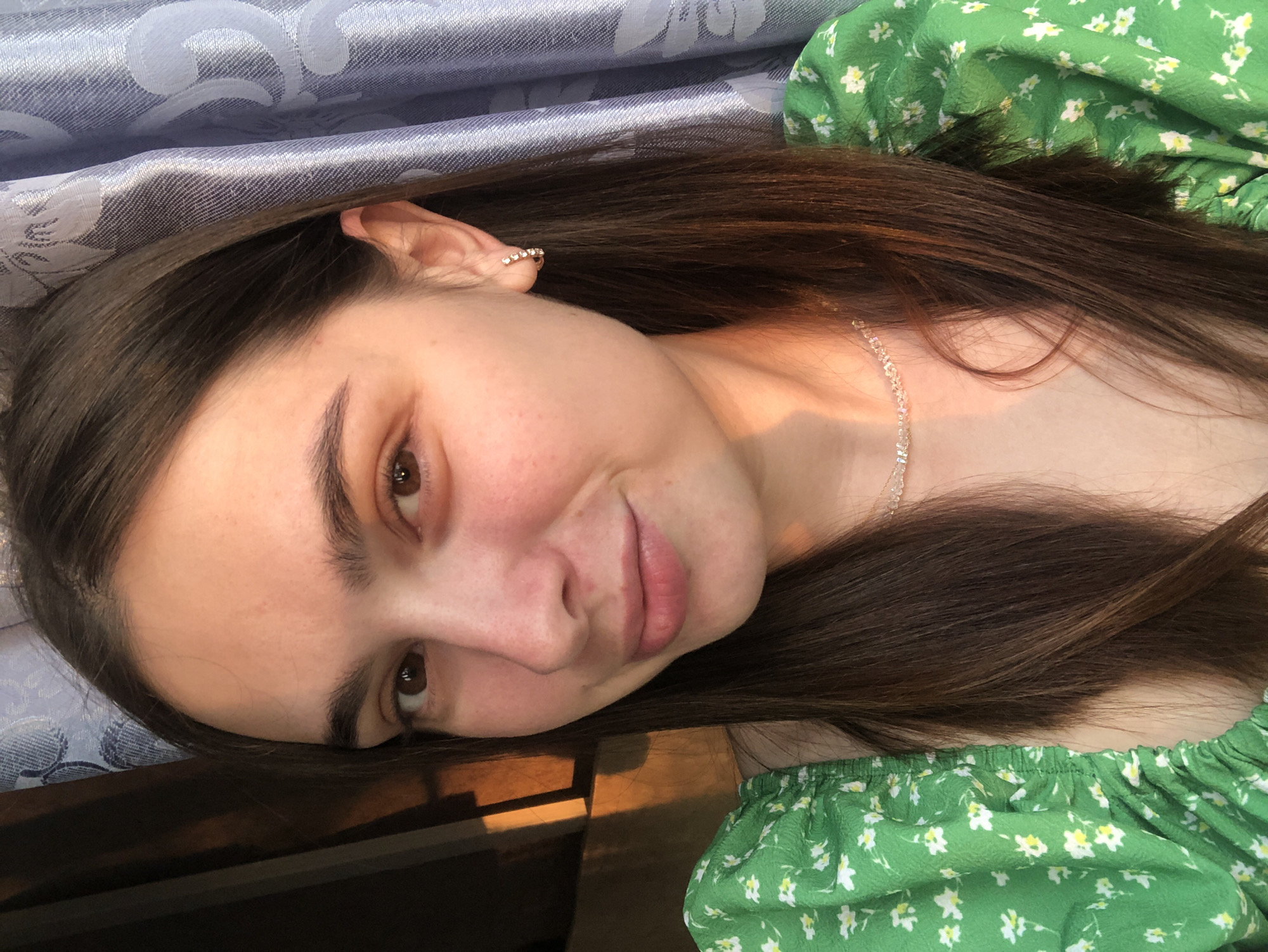 A young woman with long, dark brown hair is photographed from the chest up, facing the camera. She is wearing a green, floral-patterned top with puffed sleeves and a delicate, sparkly necklace. A small, hoop earring with tiny stones is visible on her right ear. The lighting suggests it is either sunrise or sunset, casting warm, golden light on her face and the left side of the image, where a shadowy structure is visible. To the right, a textured, light blue curtain with a floral pattern is partially visible. The background is out of focus, with the woman's face and upper body being the primary subjects. The image appears to be taken indoors, with natural light coming from a window or opening. There is no information to determine the location in Chaikovsky, Russia.