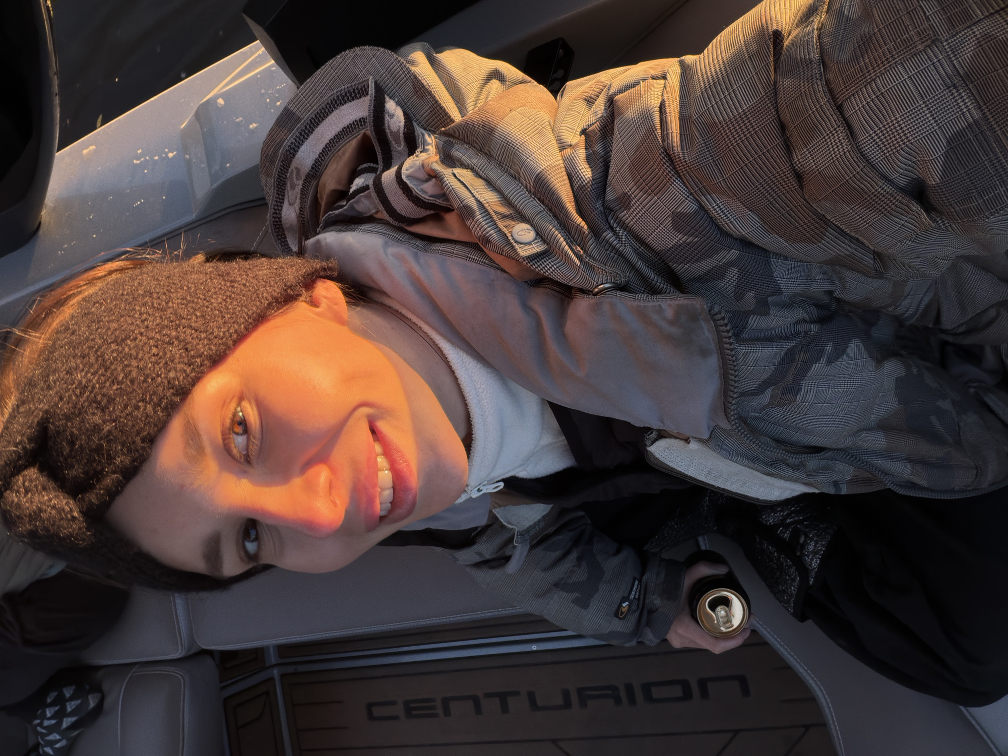 A woman wearing a dark beanie and a plaid hooded jacket sits inside a Centurion boat. She holds a gold-colored can in her gloved hand. The setting sun casts an orange glow on her face. The location is possibly near Терехово, Russia.