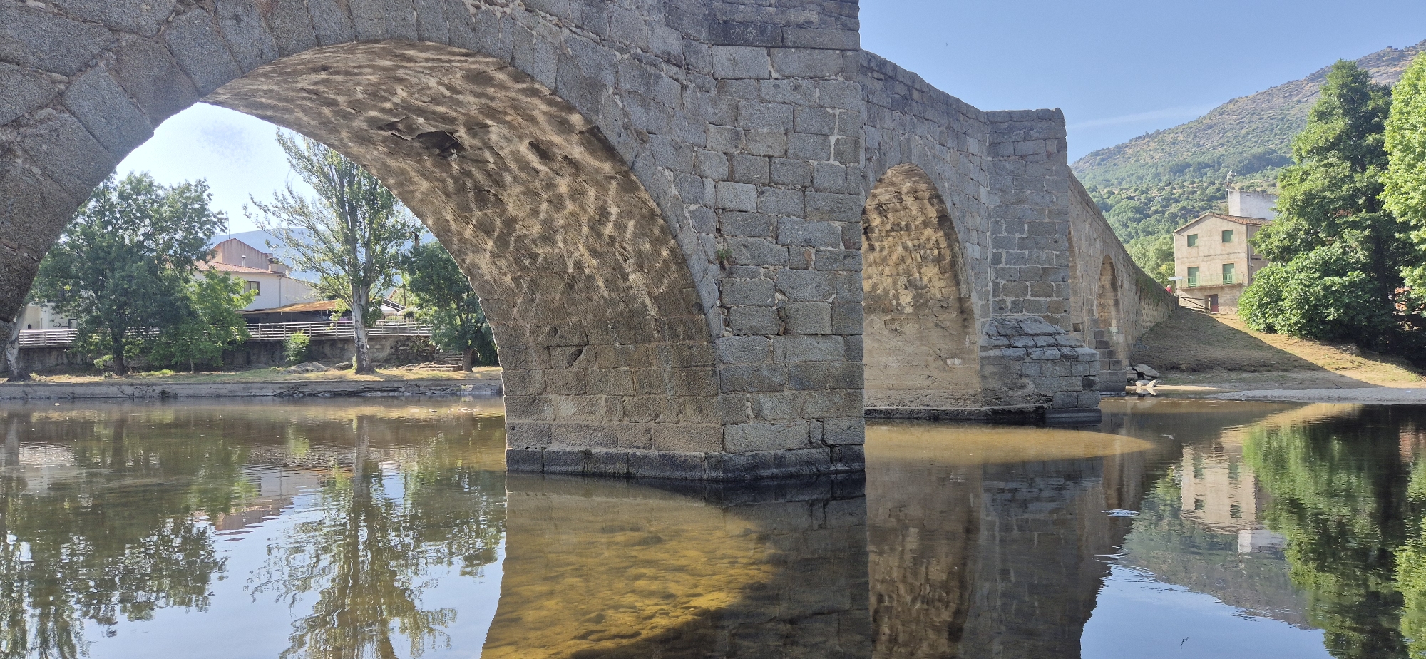 This image showcases a stone bridge spanning a body of water, likely a river, under a clear blue sky. The bridge exhibits multiple arches, with the foreground arch being the most prominent. The stonework of the bridge appears aged, with visible texture and some imperfections. Sunlight casts dappled patterns on the underside of the arch.
On the far bank of the water, a gentle slope rises, covered in green vegetation and trees. A light-colored building with a reddish-brown roof and green window shutters is visible amidst the trees, situated on this slope. Beyond the building, rolling hills covered in denser foliage recede into the background.
The water in the foreground is calm and reflects the bridge, trees, and the building. The shallow areas of the water reveal the sandy or rocky bottom. The scene suggests a peaceful, rural or semi-rural setting, possibly in a European locale, given the architectural style of the bridge and the building. There are no people visible in the image.