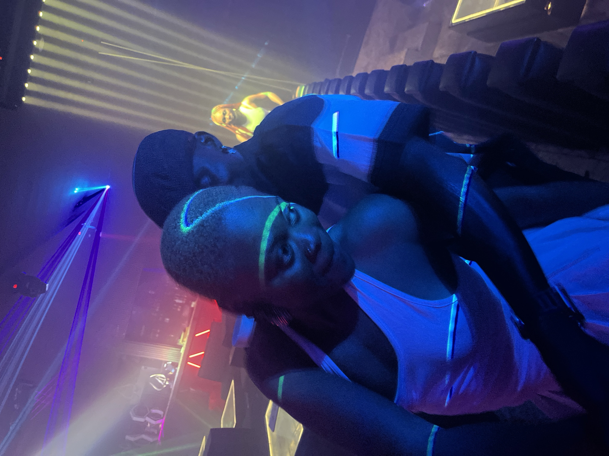 A close-up, slightly blurred, eye-level shot captures two individuals in a dimly lit, energetic indoor venue, possibly a club, in the city of Howayi, Nigeria. The scene is bathed in vibrant purple and blue neon lighting, with streaks of yellow light emanating from overhead fixtures, suggesting a dynamic atmosphere. In the foreground, a woman with dark skin and neon green makeup accentuating her eyes and forehead, wearing a white tank top, is embracing a man who is mostly obscured. He is wearing a black cap. Their bodies are pressed close together, indicating an intimate interaction. In the background, out of focus, another woman with blonde hair can be seen standing. The environment appears to be a seating area with tiered, dark structures that resemble stadium seating. Lasers cut through the smoky air, creating a visual spectacle. The overall impression is one of a lively night out, with a focus on the immediate interaction between the couple.