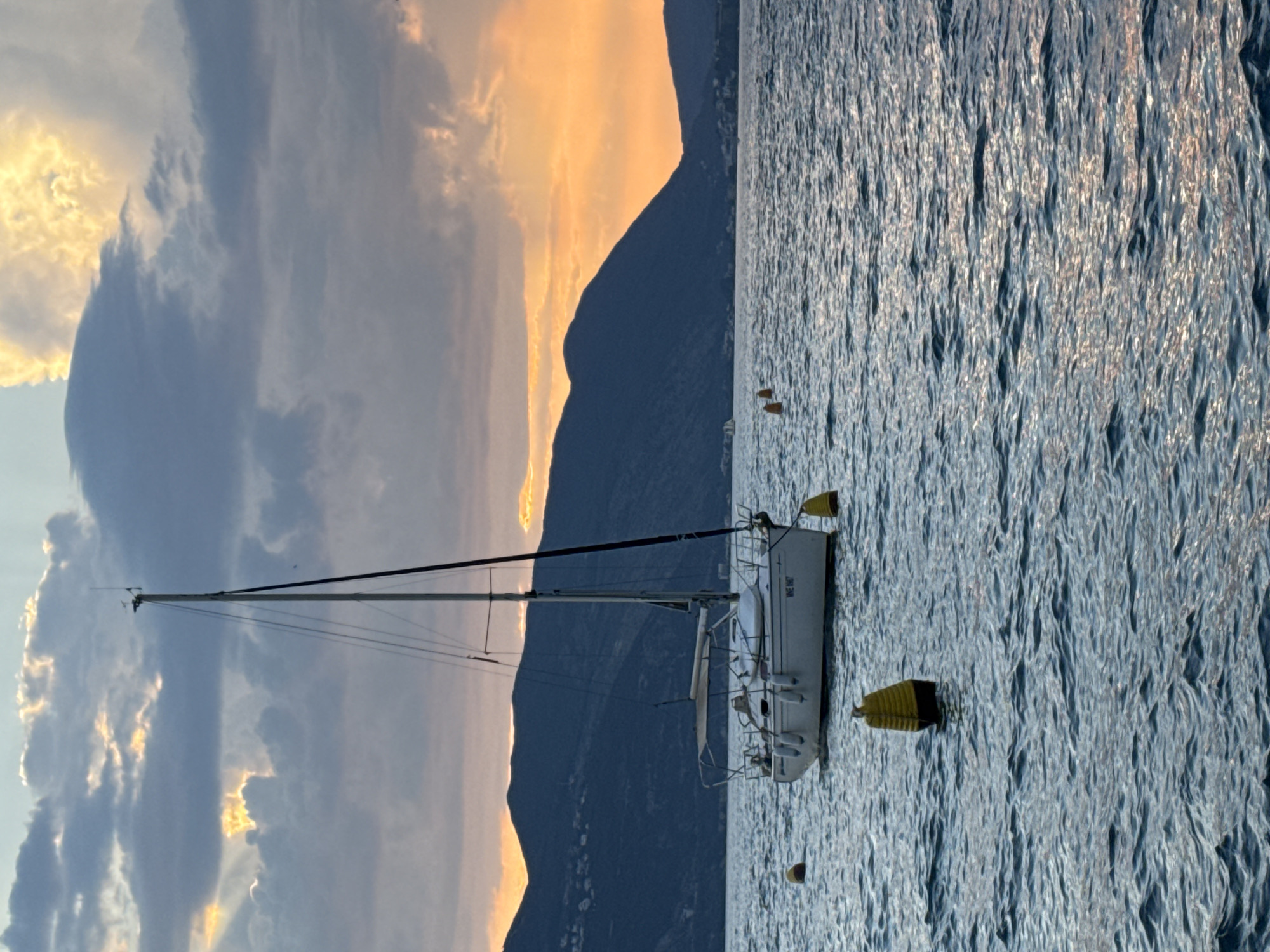 A white sailboat is anchored in the calm, blue water of a bay. The boat has a single mast and a white hull with a dark blue waterline. It is equipped with several fenders along its port side. The boat is situated in front of a line of dark, rolling hills that form the coastline. The sky above is filled with dramatic clouds, with streaks of orange and yellow light from the setting sun breaking through in places. Several yellow and black buoys are scattered in the water around the sailboat. The scene appears to be a tranquil evening on a body of water, likely a lake or a sea, with mountains in the background.
