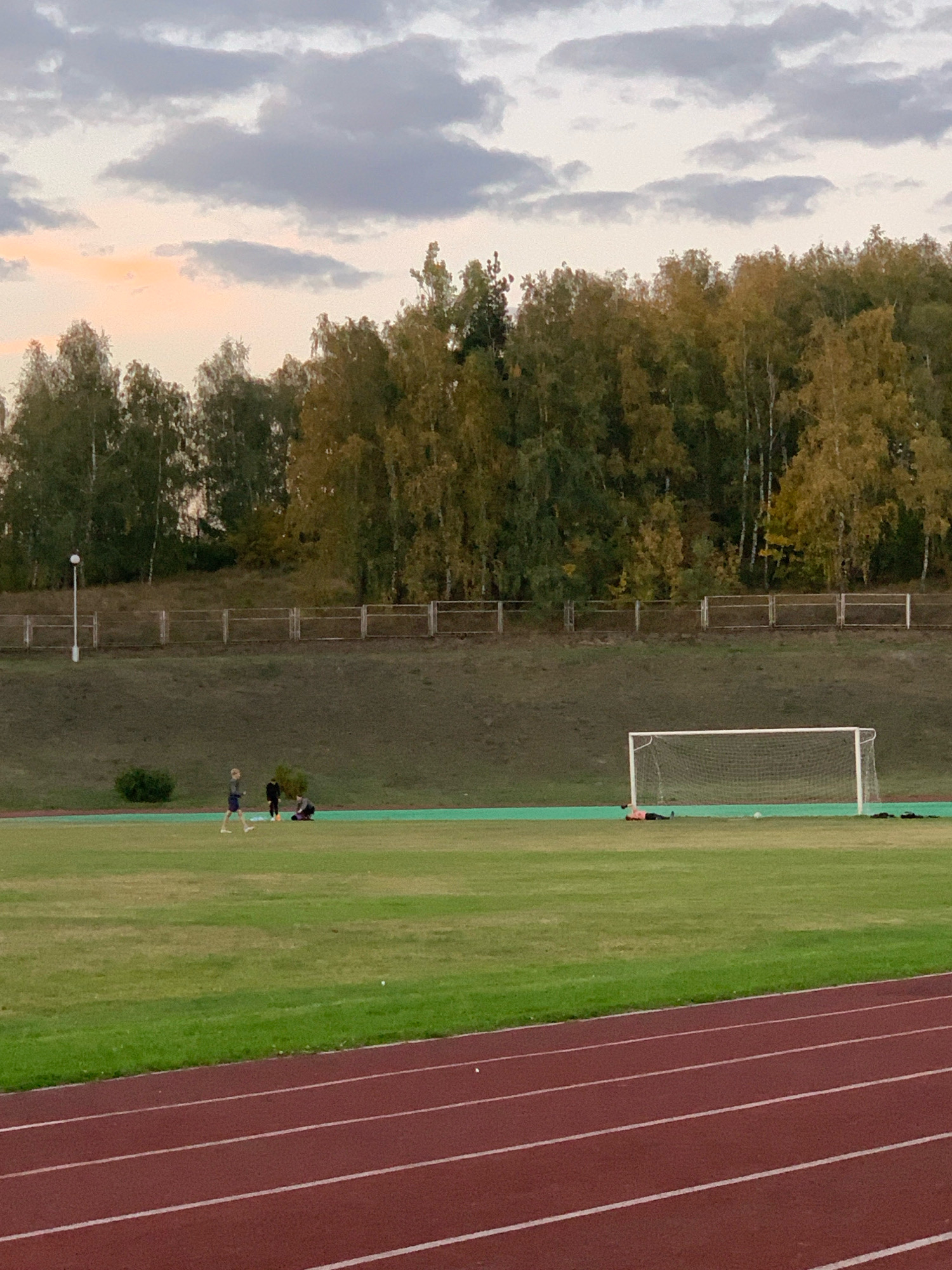 In Kapyĺ, Belarus, an outdoor sports field is depicted, with a running track in the foreground and a grassy field beyond. A soccer goal is visible on the field. In the middle distance, a group of people are present. One person, likely a child or young adult, is running. Two other individuals are seated on the ground, and another person is lying down on the field. The background features a gentle slope covered in grass, with a fence running along the top. Further back, a line of trees with autumn foliage borders the field. The sky above is overcast with some clouds.
