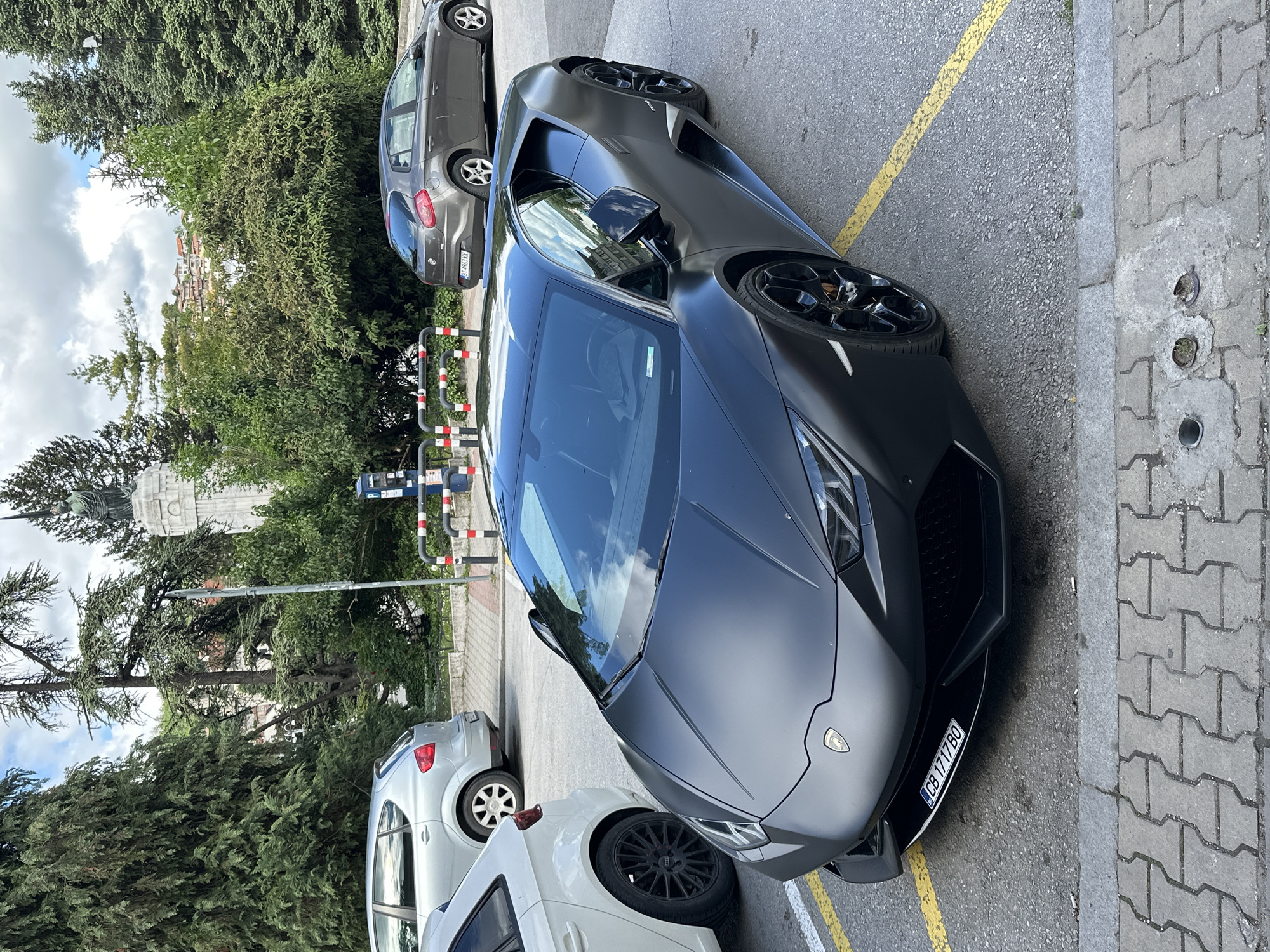 The image shows a matte black Lamborghini Huracan parked on a street in Veliko Tarnovo, Bulgaria. The car is parked next to a grey compact car and a white sedan. The license plate of the Lamborghini is visible, reading "CB 1717 BO". A yellow painted line marks the parking space. The background includes trees and a building with a monument on top. A street with red and white poles can also be seen. The street is paved with grey pavers.
