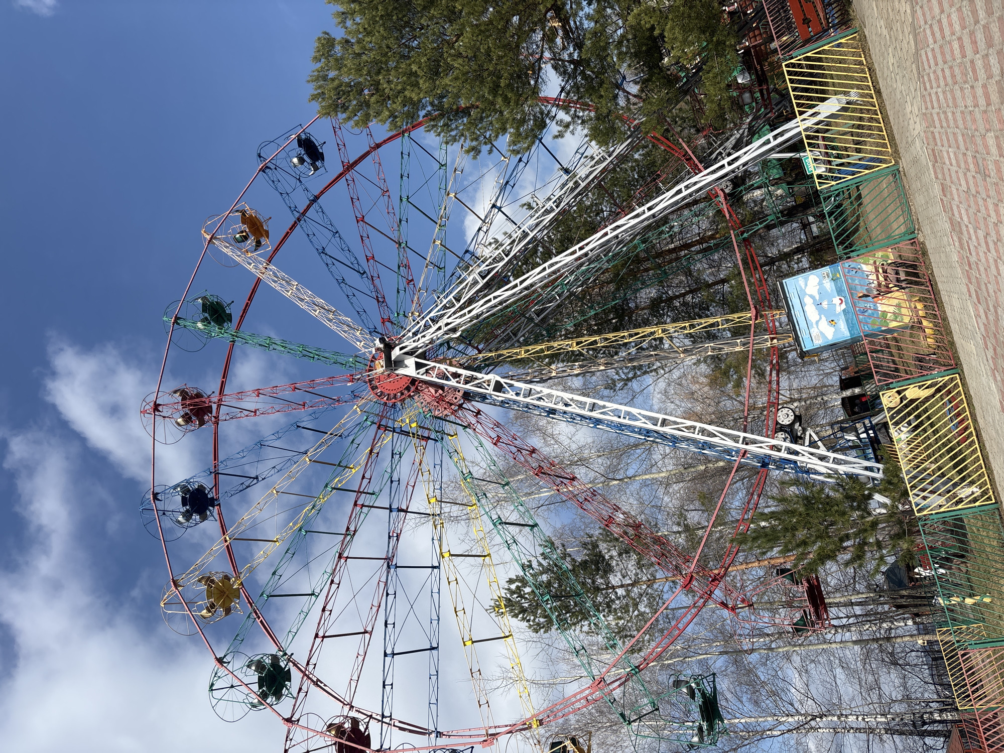 The image shows a colorful Ferris wheel, a popular amusement ride. It's located in the city of Chaikovsky, Russia. The Ferris wheel's structure is composed of red, blue, green, yellow, and white spokes, with each spoke supporting a passenger car. The sky is blue with patches of white clouds. Trees, both bare and with green foliage, are interspersed throughout the scene, partially obscuring the background. A section of patterned brick ground is visible on the right. There are no people visible.
