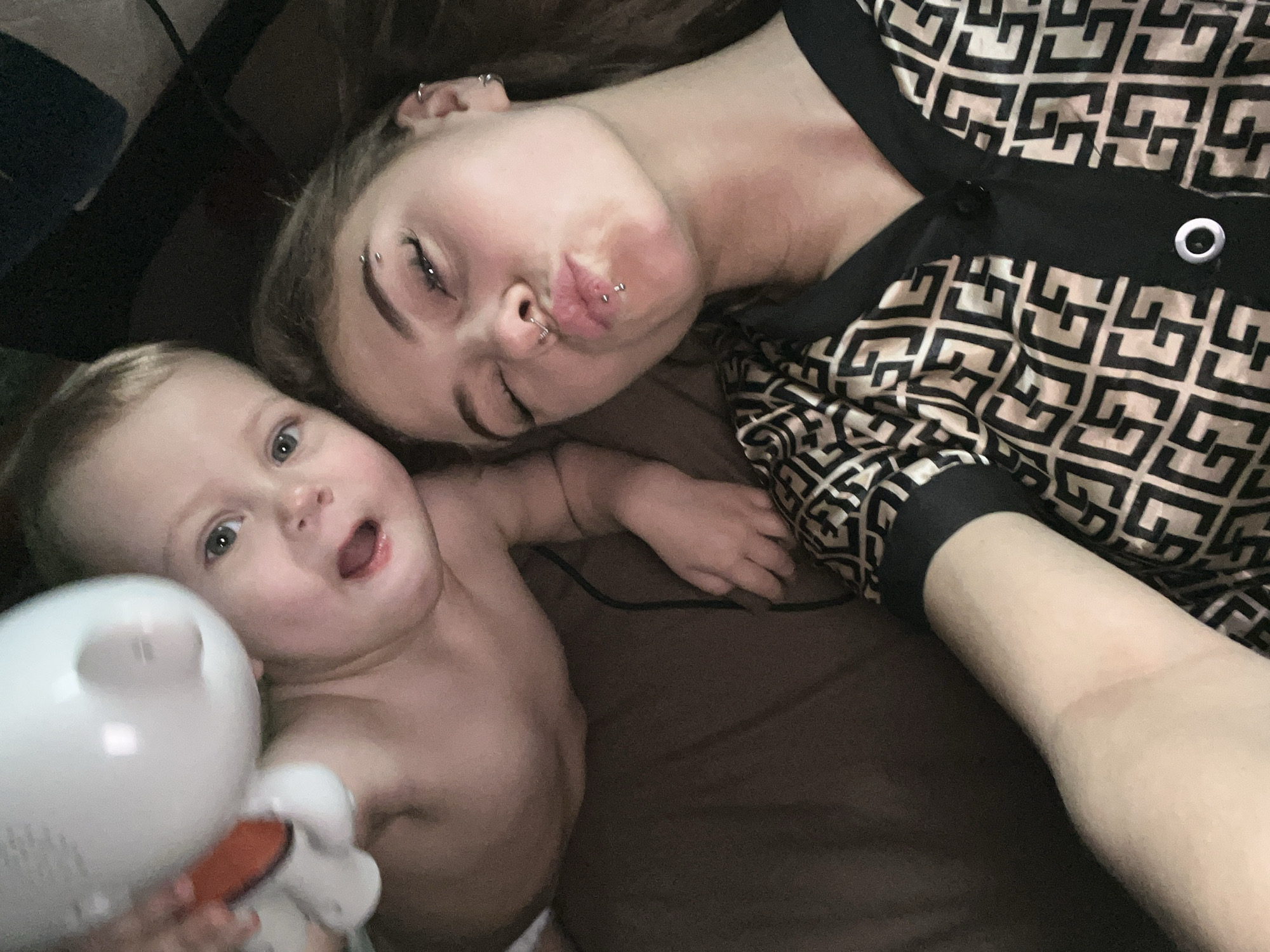A young woman with multiple piercings on her face, including eyebrow, lip, and ear piercings, is lying on a brown surface next to a baby. The woman is wearing a button-up shirt with a black and tan geometric pattern. The baby is holding a white object with an orange accent. The location is unknown and there is no information to suggest that the image was taken in the city of Фоки, Russia.