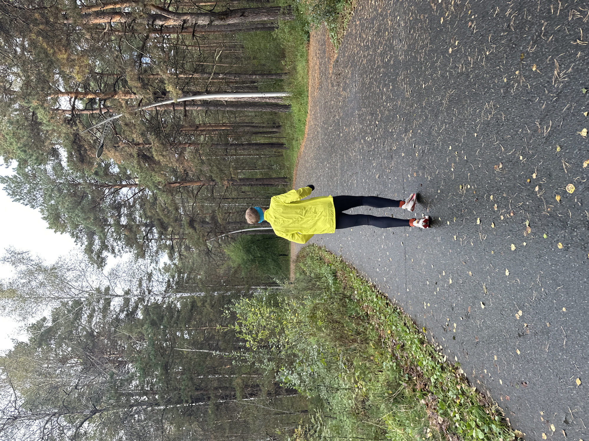 An individual is captured from behind, running on an asphalt path in a forest. The person is wearing a bright yellow jacket, black leggings, and red and white running shoes. A blue headband is visible on their head. The path is wet and scattered with fallen leaves, indicating an autumn setting. Tall pine trees and deciduous trees with sparse foliage line both sides of the path, creating a natural, wooded environment. The sky is overcast, contributing to the subdued lighting of the scene.