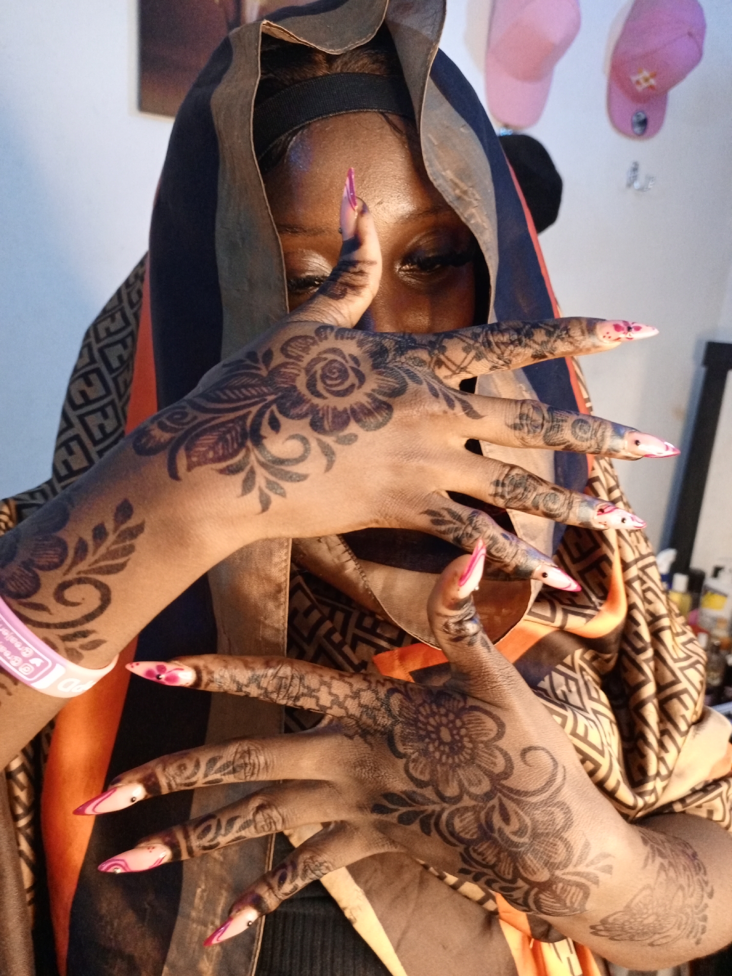 A medium shot depicts a dark-skinned woman with henna designs on her hands and fingers. She is wearing a headscarf with geometric patterns in black, brown, and orange. Her hands are positioned in front of her face, displaying floral henna patterns. She has long, pointed nails with a white base and pink tips. The background shows a glimpse of a room with pink and white baseball caps hanging on the wall.