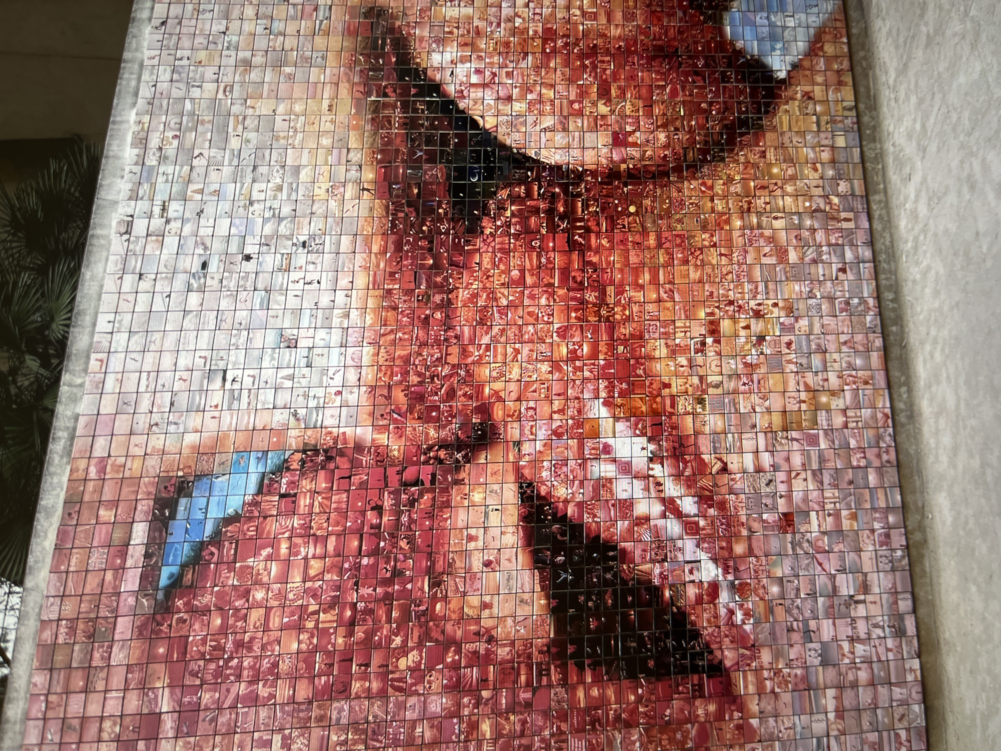 The image depicts a mosaic mural composed of small, square tiles. Each tile contains a different image, and collectively they form a larger picture of a person. The individual appears to be wearing a dark garment with a high collar, and the overall color palette leans towards reds, oranges, and browns. The mosaic is mounted on a wall, presumably indoors, with a neutral-colored background. The location of the mosaic within a specific city and state is unknown.