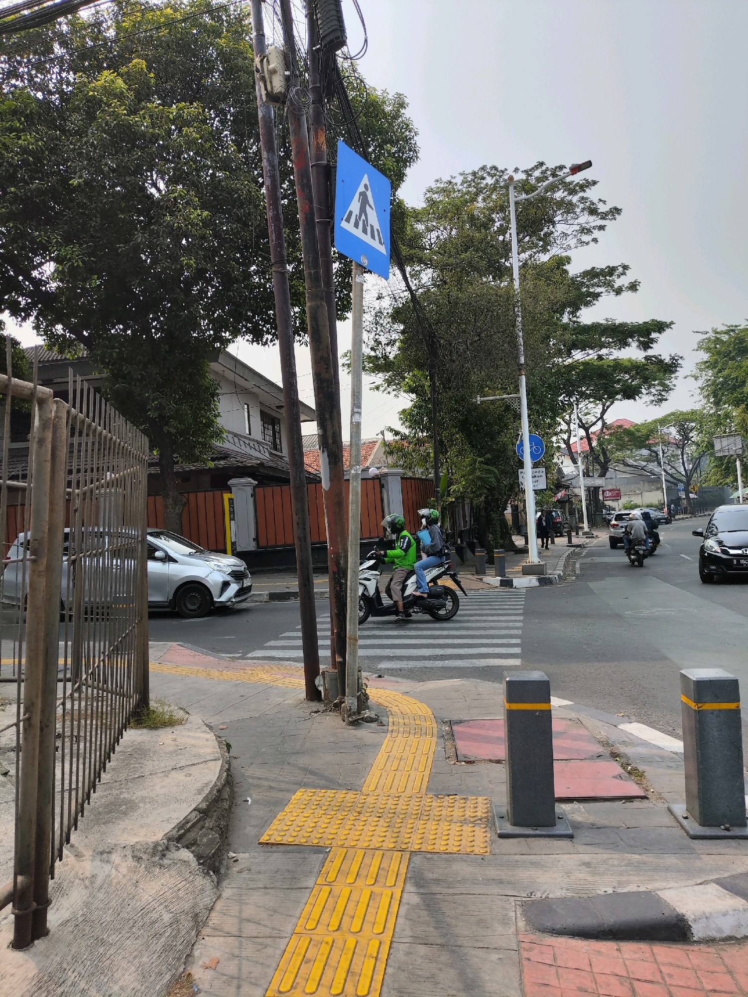 The image captures a street scene in South Jakarta, Indonesia, specifically at a pedestrian crossing. In the foreground, a textured yellow tactile paving guides pedestrians, transitioning to a concrete sidewalk with a decorative tile border. A metal fence runs along the left side of the sidewalk.
Across the street, a silver car is parked near a building with dark red walls and a white upper story. A zebra crossing is clearly marked on the asphalt road. Two individuals on a white motorcycle, both wearing green jackets and helmets, are in the process of crossing the street.
Further down the road, other vehicles, including motorcycles and cars, are visible. Traffic lights and street signs are present, indicating a regulated intersection. The environment is characterized by lush green trees bordering the road and the sky overhead is overcast. The overall impression is that of a typical urban street in Indonesia, with a focus on pedestrian safety at the crossing.