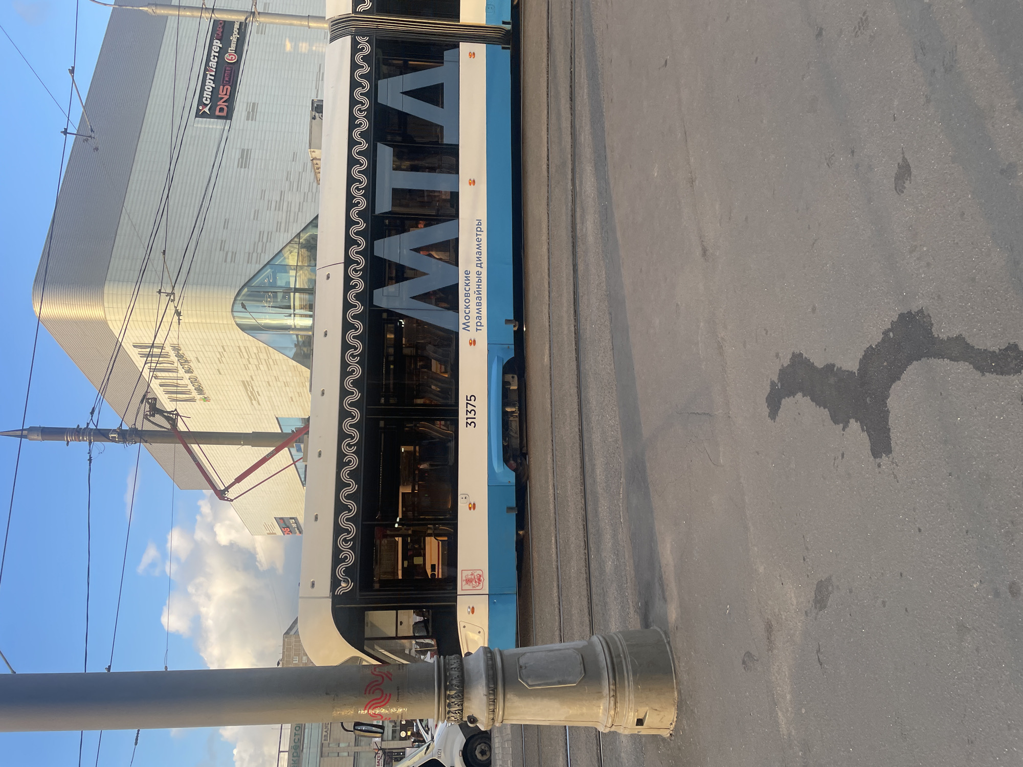 The image shows a tram with the text "МТА Московские трамвайные диаметры" and the number "31375" on its side. The tram is white and blue, with a wavy black and white pattern along the top. Large letters spelling "MTA" are visible through the windows. In the background, there is a modern building with a white and grey facade, and a blue sky with clouds. Power lines for the tram are visible. The scene is set on a street with asphalt pavement and tram tracks. There are no people clearly visible in the image. The building in the background appears to be a commercial or public building, possibly a shopping center, with signage that includes "СПОРТМАСТЕР" and "DNS". The overall impression is an urban scene in Moscow, Russia.