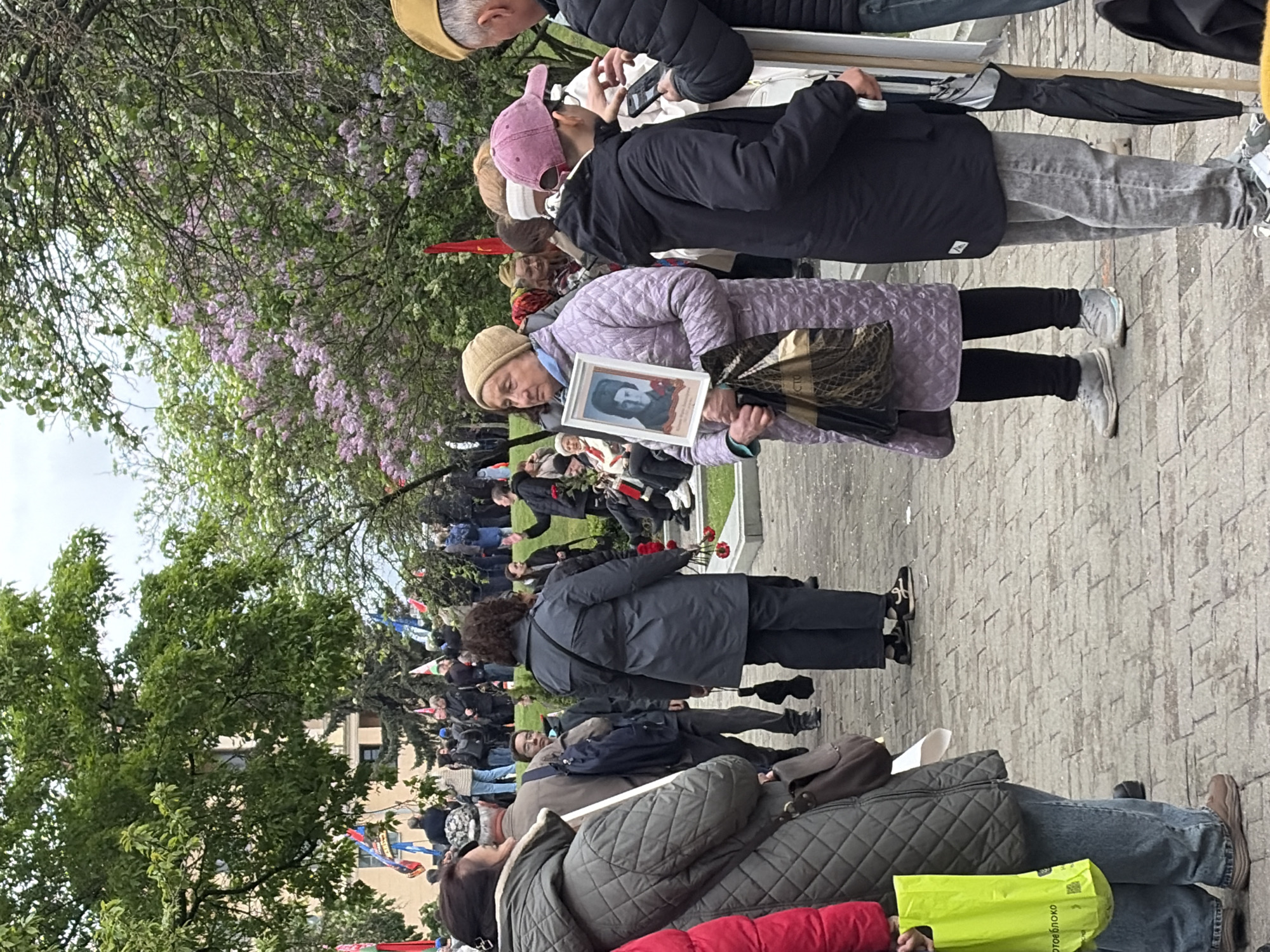 A horizontal eye-level shot captures a crowd of people gathered in a city park, identified as being in Unknown, Unknown. The environment includes flowering trees, adding a springtime atmosphere. A gray-bricked walkway is visible in the foreground and extending into the background. The focus of the shot centers around an older woman dressed in a light purple quilted coat and a tan beanie. She stands out as she holds a framed portrait of a person, presumably a deceased relative.
Surrounding the woman, other individuals are either standing or walking, some of whom are carrying items such as bags or umbrellas. A person with a pink baseball cap is visible behind her, seemingly taking a photo with a smartphone or camera. Several flags are visible further back in the crowd, potentially indicating a political or commemorative gathering. Towards the back, people are placing red flowers at a memorial.