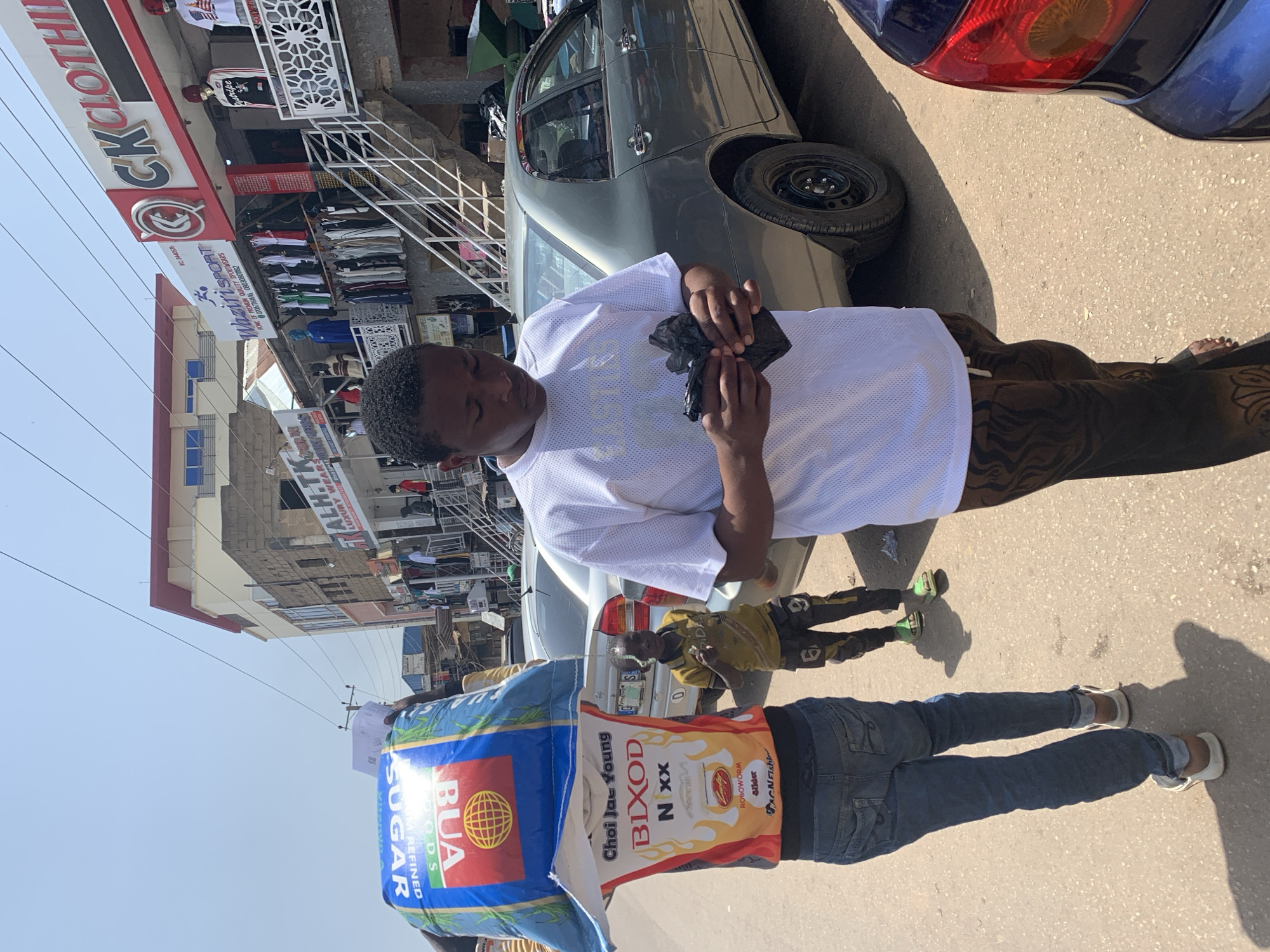 The image shows a street scene in Jos, Nigeria, with several people and vehicles. In the foreground, a young man wearing a white t-shirt and patterned pants is looking at something in his hands. To his left, another person is carrying a large blue and white bag of sugar on their head. This person is wearing a t-shirt with graphics and text, including "BUA" and "SUGAR". Behind them, a young boy in a yellow t-shirt and dark pants with yellow accents stands with his hand raised. A grey sedan is parked to the right of the frame, and a dark blue car is partially visible on the right edge. The background features buildings with shops, including "CK CLOTHING" and "WAZIRI SPORT", with clothing displayed on mannequins and racks. The street is paved with asphalt and shows signs of wear. Power lines are visible overhead, and the sky is clear and bright, indicating a sunny day. The overall scene depicts a bustling commercial area.