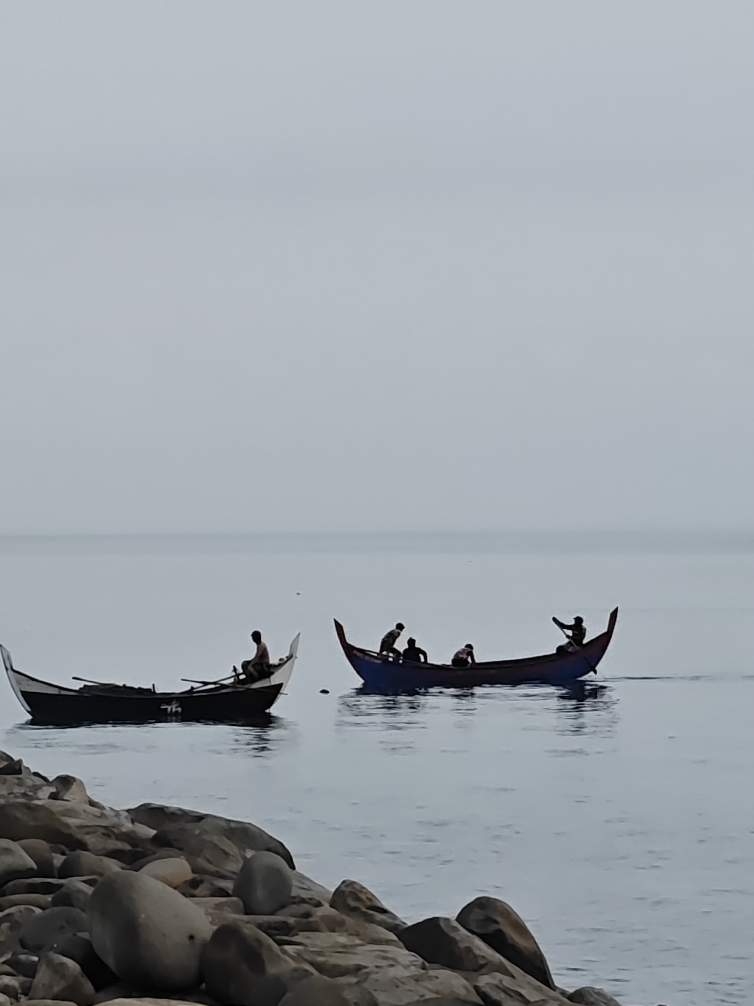 This media file captures a serene coastal scene in Lhokseumawe, Indonesia, featuring two traditional fishing boats on calm, grey-blue water. The sky is uniformly light grey, indicating an overcast day or possibly early morning/late afternoon, with a faint horizon line.
In the foreground, large, dark grey and brown rocks line the water's edge. Beyond them, two slender, pointed-bow boats (likely perahu) are visible.
The boat on the left has a dark hull and a lighter, possibly white, upper section at its bow and stern. One person, a silhouette, is seated within, holding what appears to be a long oar or paddle. A distinctive white, stylized symbol, possibly the letters "TH" or a similar emblem, is visible on the side of this boat.
To the right, a second boat, larger and more distinctly colored with a blue hull and hints of red interior or trim, carries at least four people. One person at the stern is actively paddling, creating a subtle ripple behind the boat, suggesting it is in motion. The other individuals are silhouetted against the water, their exact activities unclear but suggesting a collective effort, possibly heading out for fishing or returning.
The water is still and reflective, mirroring the soft, diffused light from the sky. The overall atmosphere is quiet and tranquil, depicting a moment of daily life or work on the water. No distinct text is discernible other than the stylized symbol on the left boat.