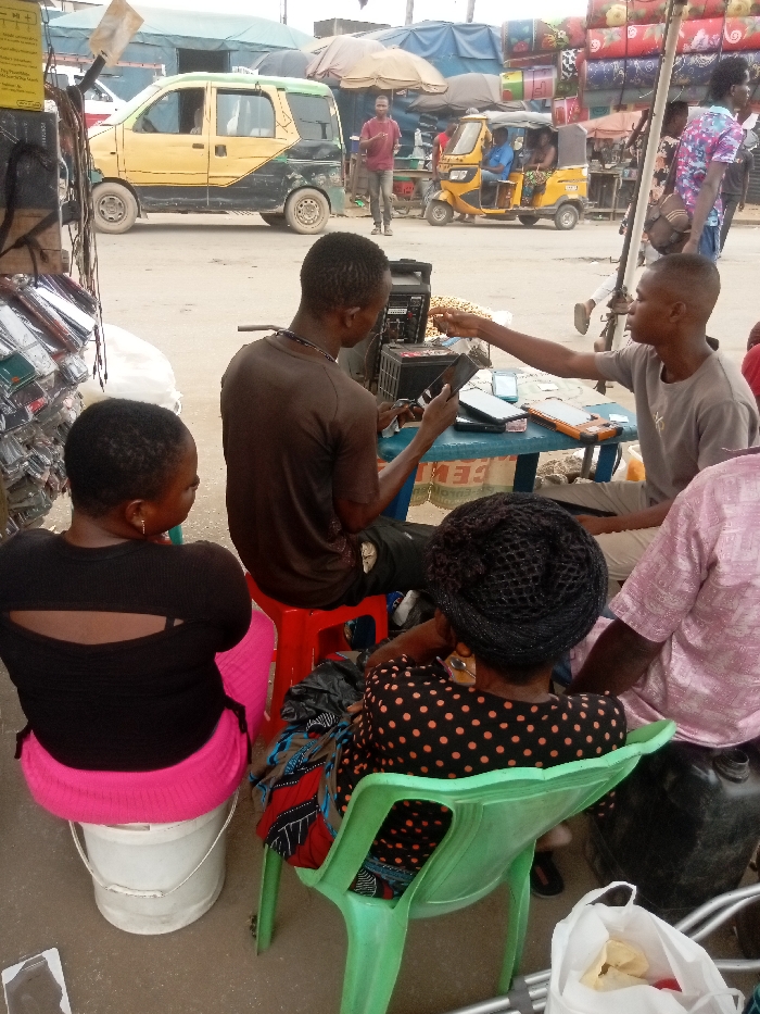 The image captures an active outdoor market scene in Idemili, Nigeria. In the immediate foreground, five individuals are situated around a low, rectangular blue plastic table with white legs.
Two males are centrally engaged at the table. One male, with his back to the camera, wears a dark brown shirt and sits on a red plastic stool. He holds a dark mobile phone in his left hand and appears to be operating a black portable power generator or inverter, equipped with a battery and visible wiring, positioned to his left on the ground. Opposite him, a second male in a light gray t-shirt extends his right arm, presenting a dark rectangular object, consistent with a mobile phone or tablet, towards the first male. Various other mobile phones and electronic devices are displayed on the table, alongside a small pile of peanuts.
Three other individuals are seated around this central interaction. To the far left, a female wearing a black top with shoulder cutouts and a vibrant pink skirt is seated on an inverted white plastic bucket. To the right of the male operating the power source, a female in a black polka-dot top and a black mesh head covering sits on a light green plastic chair. Partially visible on the far right, a fifth person wears a light pink patterned garment. On the far left edge of the frame, a vertical display rack holds numerous transparently packaged phone cases or accessories.
The background shows a busy, unpaved street. A two-tone yellow and black van is parked or moving slowly on the left side of the thoroughfare. Further down the street, a yellow motorized rickshaw (keke napep) carries passengers. Several pedestrians are visible walking throughout the scene. The street is bordered by informal market stalls constructed with various tarpaulin coverings (blue, brown, green), with one prominent stall on the right displaying a tall stack of colorful, patterned cushions or mattresses.