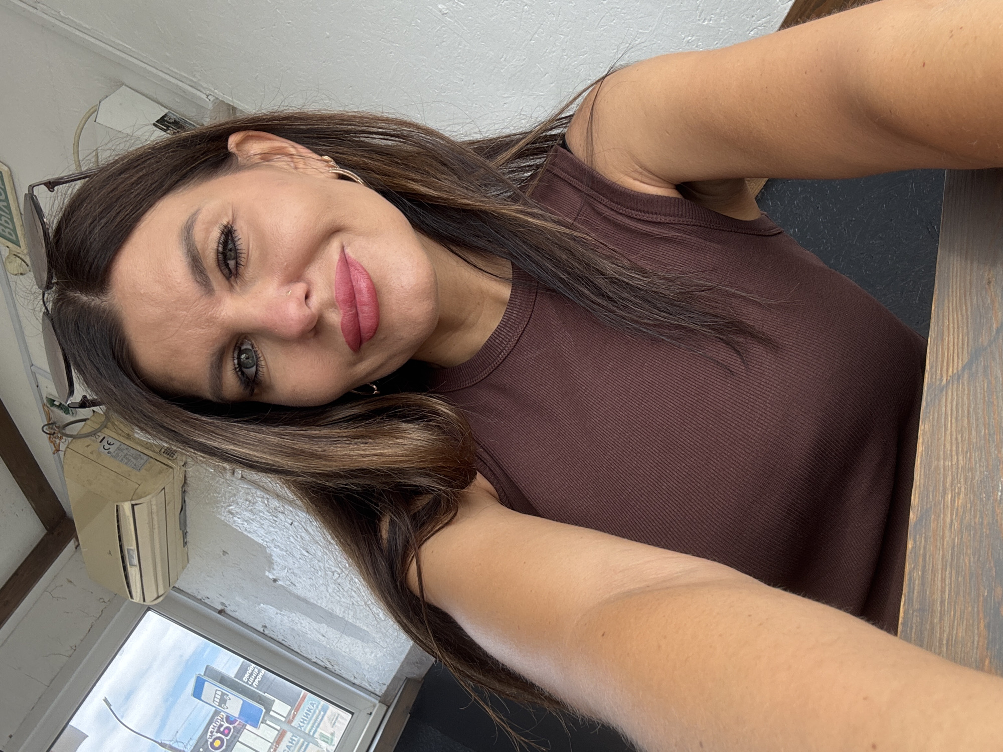 A medium shot selfie captures a woman in her early 30s with long, dark brown hair and blue-green eyes, facing the camera. She is wearing a brown, sleeveless, ribbed top. Her makeup includes eyeliner, mascara, and pink lipstick. She has a small nose piercing and is wearing a hoop earring in her right ear. Her left arm is extended towards the camera, and her right arm is bent, with her elbow resting on a wooden surface.
The background suggests an indoor setting, possibly a cafe or restaurant. To the left, a window reveals an outdoor scene with a cloudy sky, a street lamp, and a blue bus stop sign with text in Russian. The wall behind her is painted white and shows signs of wear. An off-white, outdated air conditioning unit is mounted on the wall above and to the left of her head. A sign with green lettering in Russian, partially obscured by her hair, is visible on the ceiling. The overall lighting is natural and appears to be coming from the window. The composition is informal, typical of a casual selfie.
The bus stop sign visible in the background, with Cyrillic script and the characteristic blue and white colors, strongly suggests the location is within Russia. Based on the presence of a bus stop sign and the general visual cues, it is plausible this image was taken in the city of Chaikovsky, Russia, given its known infrastructure and urban environment.