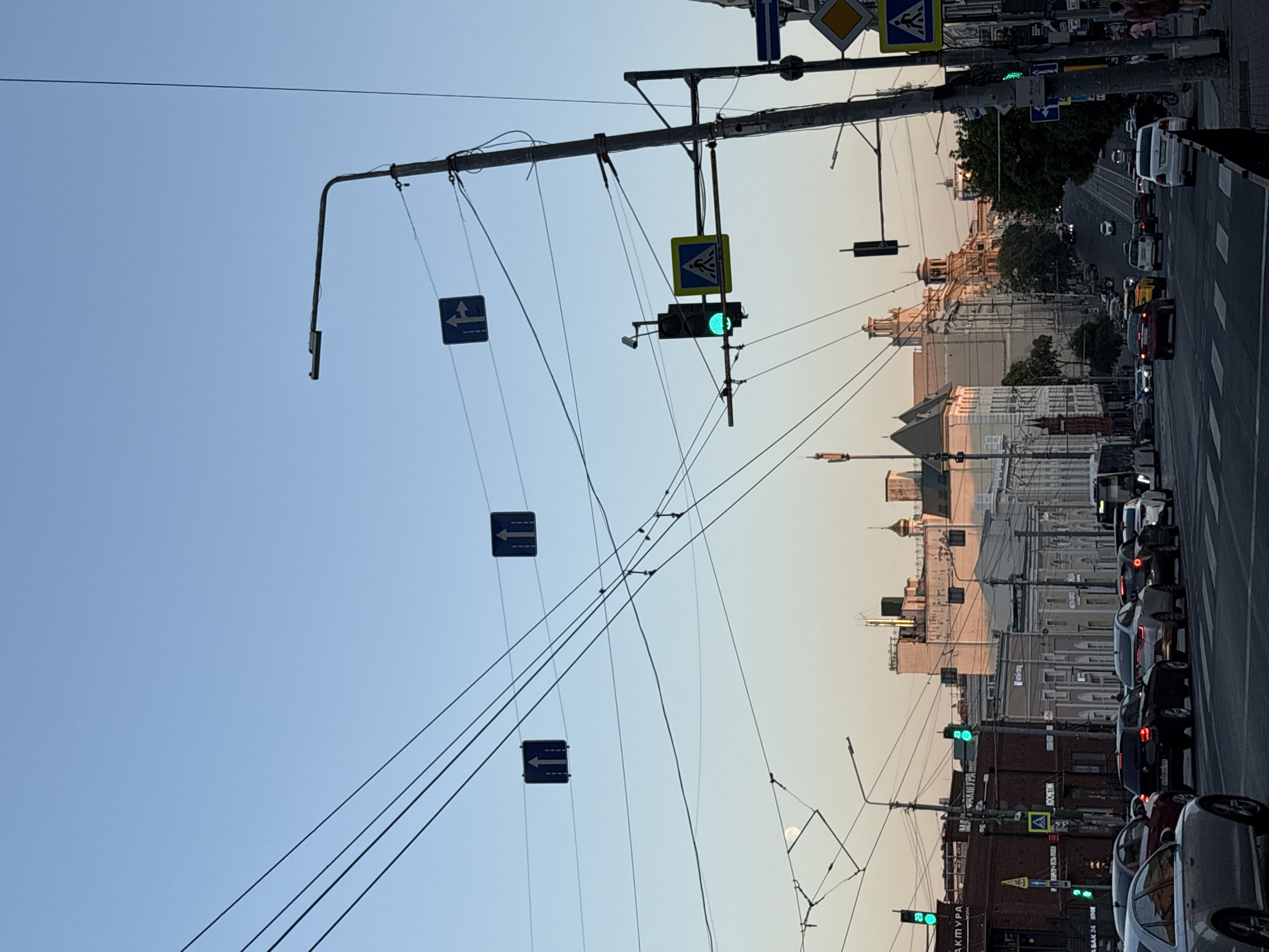 The image depicts a street scene in Rostov-on-Don, Russia, during the late afternoon or early evening. The sky is clear and a pale blue, transitioning to a warmer hue near the horizon. A complex network of overhead wires, likely for trolleybuses or trams, crisscrosses the frame, casting faint shadows.
Traffic is moderate, with cars lined up at what appears to be an intersection controlled by traffic lights. Several traffic lights are visible, with at least one showing a green signal. Road signs, including directional arrows and a pedestrian crossing symbol, are mounted on poles and suspended from the wires.
Buildings of various architectural styles line both sides of the street. On the left, a building with a brick facade displays a sign with Cyrillic lettering, possibly indicating a business. Further down the street, older, multi-story buildings with decorative elements are visible. On the right, modern streetlights and utility poles stand alongside the buildings.
In the foreground, the road surface and pedestrian crossings are clearly visible. A few pedestrians can be seen on the sidewalk on the right side of the image. The overall impression is a bustling urban environment captured during a transitionary time of day.