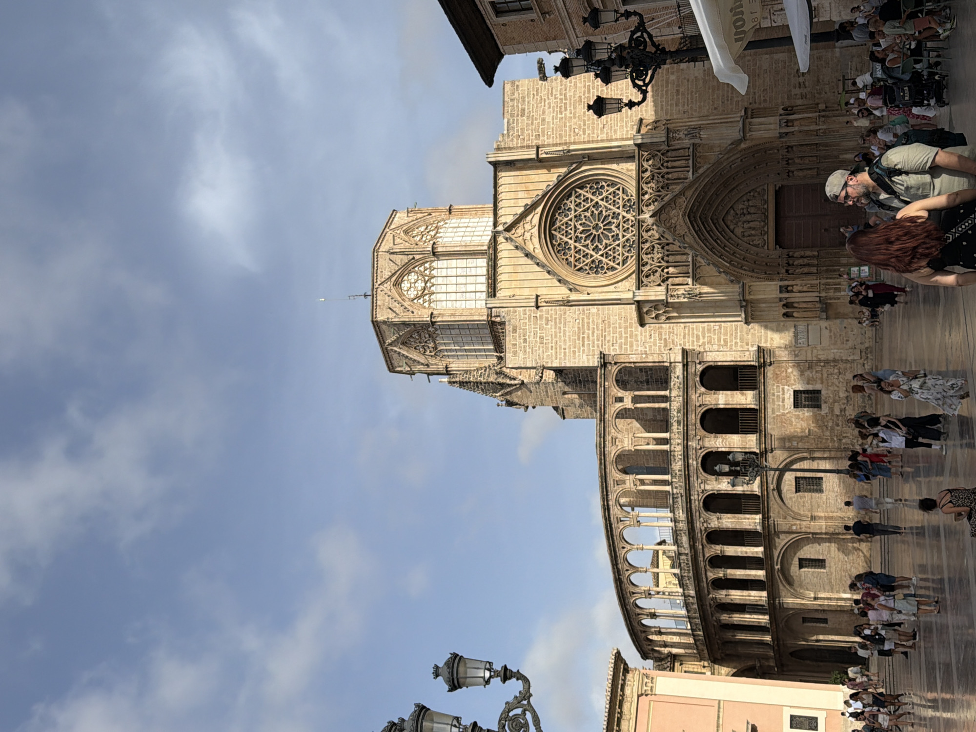 This is a vertical, eye-level, outdoor photograph taken on a bright, partly cloudy day. The scene depicts a large, ornate gothic-style cathedral with a prominent rose window above a grand arched entrance. To the left of the cathedral, there is a building with a semicircular facade featuring a series of arches supported by columns. In the foreground, a paved plaza is populated with numerous people engaged in various activities, including walking, standing, and sitting at outdoor tables. Two vintage-style street lamps with multiple lights are visible on the left and right sides of the image. The sky above is blue with scattered white clouds.