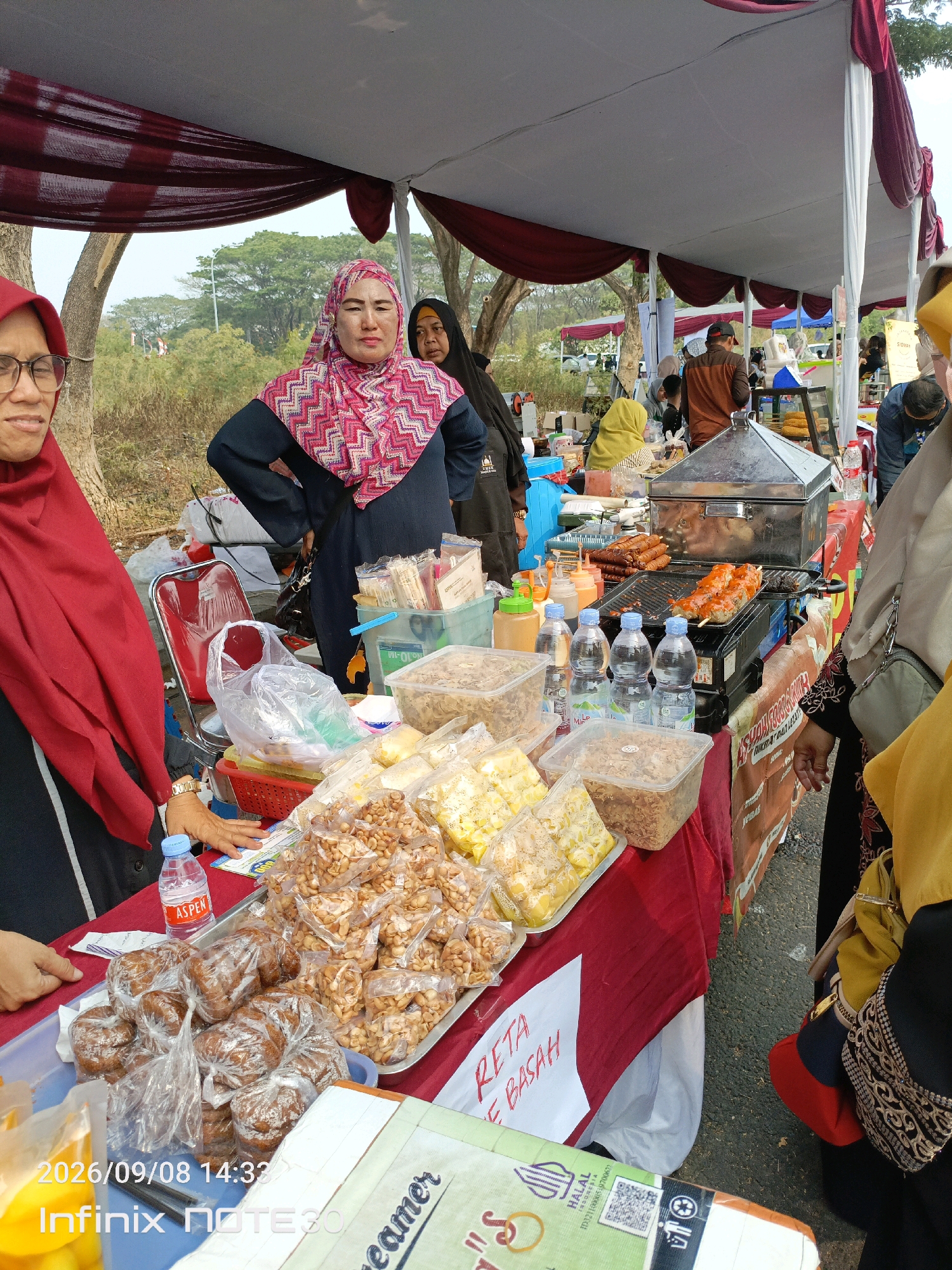 The image depicts an outdoor food stall, likely in a market or event setting in East Jakarta, Indonesia. The stall is covered by a large white and maroon tent. Several individuals are visible, primarily women wearing hijabs. In the foreground, a woman in a red hijab and glasses is on the left, and another woman in a pink and white patterned hijab is in the center. Several tables display a variety of food items, including packaged snacks like peanuts and what appear to be fried pastries, as well as grilled skewers of meat on a portable grill. Bottles of water are also present on the tables. The background shows more stalls and people, suggesting a bustling marketplace environment. The ground appears to be asphalt or a similar paved surface. The overall scene conveys a sense of a community gathering or a local market day.