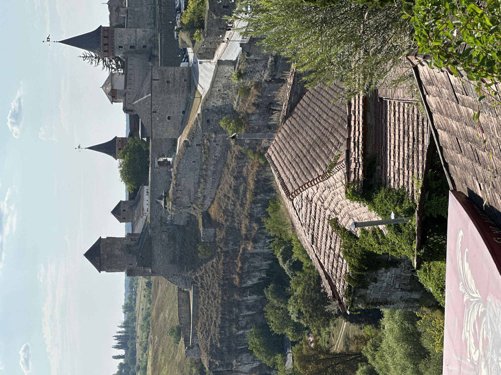This is a high-angle, long-shot photograph of the Kamianets-Podilskyi Castle in Kamianets-Podilskyi, Ukraine, on a sunny day with a partly cloudy sky. In the foreground, two tiled roofs of houses are visible, one covered in ivy. Beyond these, the imposing stone walls and towers of the ancient castle complex dominate the middle ground. The castle is situated on a rocky cliff face, with a road winding through it, where several people are seen walking. The surrounding landscape consists of rolling hills, dry grass, and scattered trees. In the distance, more trees and a hazy horizon are visible. The architecture of the castle features prominent round towers with conical roofs, stone fortifications, and arched entrances. The scene conveys a sense of historical grandeur and a warm, sunny climate.