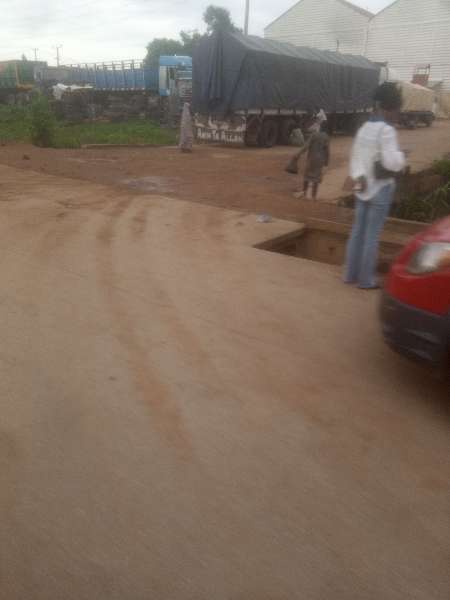 In Nassarawa, Nigeria, this image captures a dusty, outdoor scene with several trucks and individuals. A large, dark tarp-covered truck dominates the background. To its left, a blue and white truck is parked, its bed filled with stacked tires. Further to the left, another green truck is partially visible.
Several people are present. A woman in a white top and blue jeans stands in the foreground, facing away from the camera, with her legs visible. To her left and further back, a person in a grey garment is walking away from the camera, carrying an object in their right hand. Near the tarp-covered truck, a person in a patterned dress is walking towards the left of the frame. Another individual, whose clothing is indistinct, appears to be near the side of the large tarp-covered truck. The ground is a mix of dirt and some patchy wet areas. In the lower right corner, the front fender and headlight of a red car are visible. The sky is overcast.