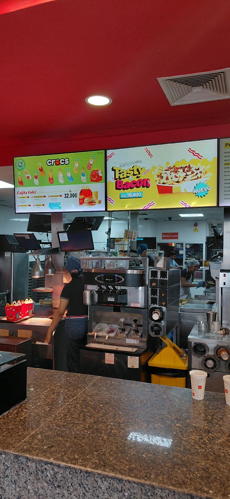 The image depicts the interior of a McDonald's restaurant, specifically at the counter area, likely in Encarnación, Paraguay, given the currency "Gs." (Guaraníes) visible on the menu boards. The scene is brightly lit, with a red ceiling and large digital menu boards.
One menu board on the left advertises "Cajita Feliz" (Happy Meal) with "Crocs" charms, priced at "Gs. 32.000". It shows various charms and a Happy Meal box with a burger and fries. The other menu board features "McPapas con Tasty & Bacon" (McFries with Tasty & Bacon), priced at "Gs. 18.000", with an image of fries topped with bacon and cheese.
In the foreground, a granite countertop is visible, with a few McDonald's branded cups. Behind the counter, kitchen operations are underway. A male employee wearing a dark shirt, blue jeans, and a dark blue cap is standing at the service counter, seemingly preparing or handing over an order. To his right, a soft-serve ice cream machine is prominent. Further back, another employee, a woman with her hair covered, is seen working at a food preparation station. A yellow trash receptacle is also visible. The overall environment suggests a busy fast-food service area.