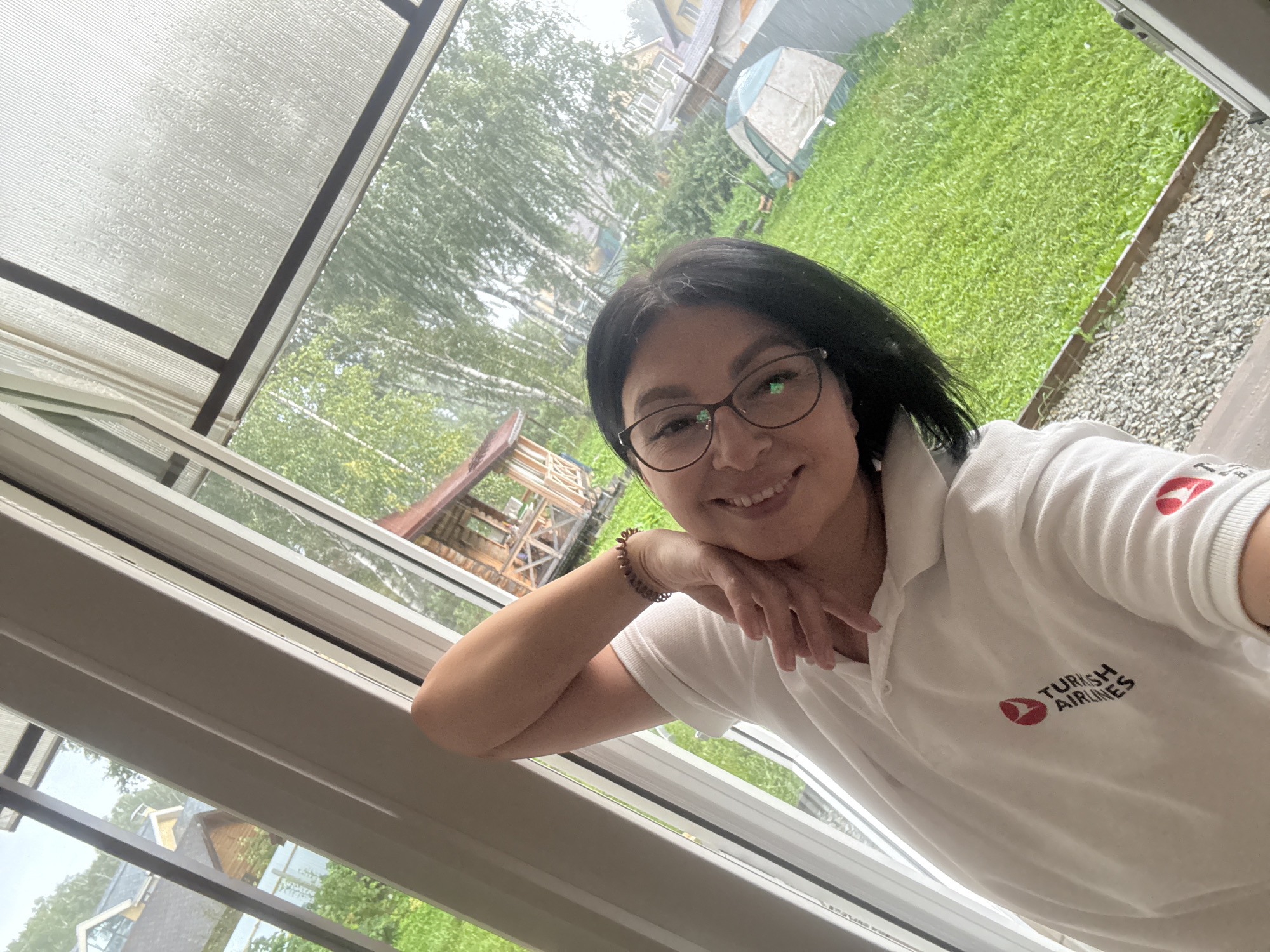 A woman with dark hair and glasses, wearing a white polo shirt with the "Turkish Airlines" logo, smiles while posing for a selfie. She is indoors, leaning against a window frame, with her left arm bent and hand near her face. Outside the window, a green lawn, a small wooden gazebo, houses, and a tent are visible under a cloudy sky. The image appears to have been taken in the village of Kremenkul', Russia.
