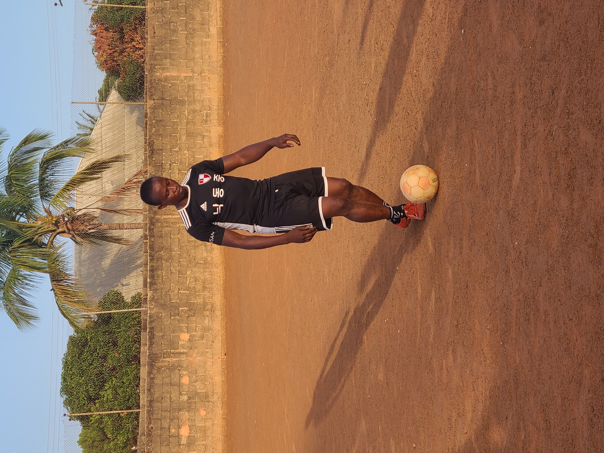 A full-length outdoor shot depicts a man poised with a soccer ball on a flat, reddish-brown dirt surface in Unknown, Unknown. The man, wearing a black athletic shirt with white markings and black shorts with a white stripe, is in a stance suggesting he is about to kick the ball. A shadow of the man is cast on the ground. Behind him, there is a concrete wall, sections of a wire fence, trees, and a glimpse of the clear blue sky.
