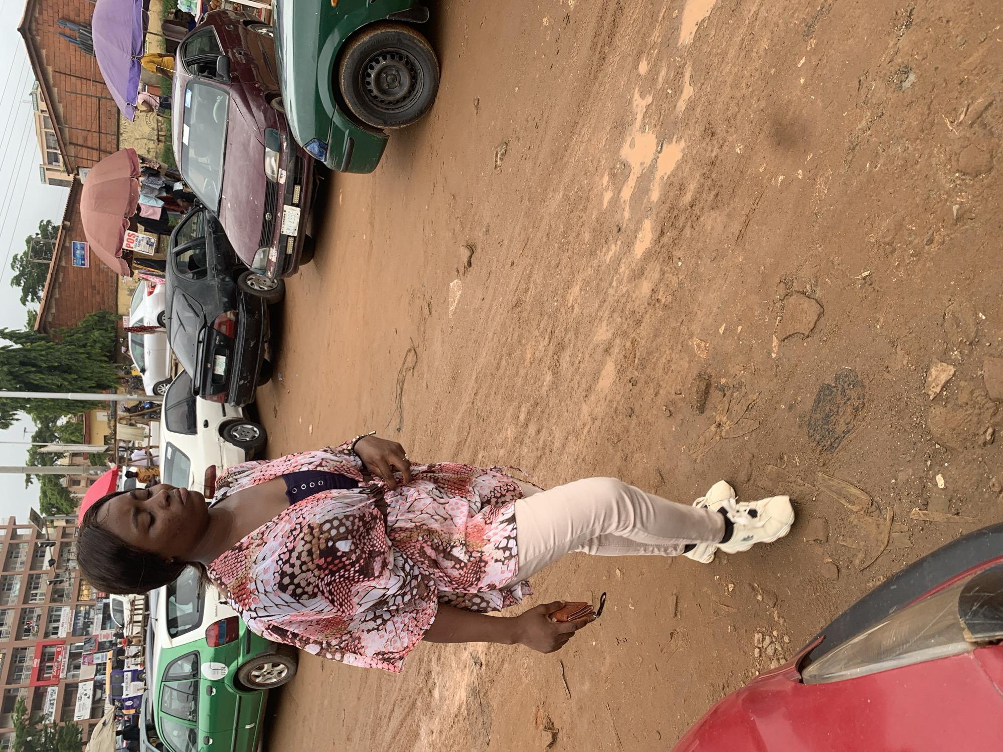 The image depicts a street scene in Abuja, Nigeria, characterized by a muddy, unpaved road surface with visible puddles, indicative of recent rainfall. Several vehicles are present, including a green hatchback, a white sedan, a black sedan, a burgundy sedan, and the front of a red vehicle in the foreground. The background shows buildings, some with signage, and market stalls covered by umbrellas.
In the foreground, a woman stands with her eyes closed, her head slightly bowed. She is wearing a patterned pink and white kimono-style top over a dark blue top, paired with light-colored trousers and white sneakers. She is holding a brown wallet or clutch in her right hand. Her posture suggests she might be praying or resting. The woman appears to be the central subject of the image. The overall atmosphere is that of a busy, possibly informal, urban environment.