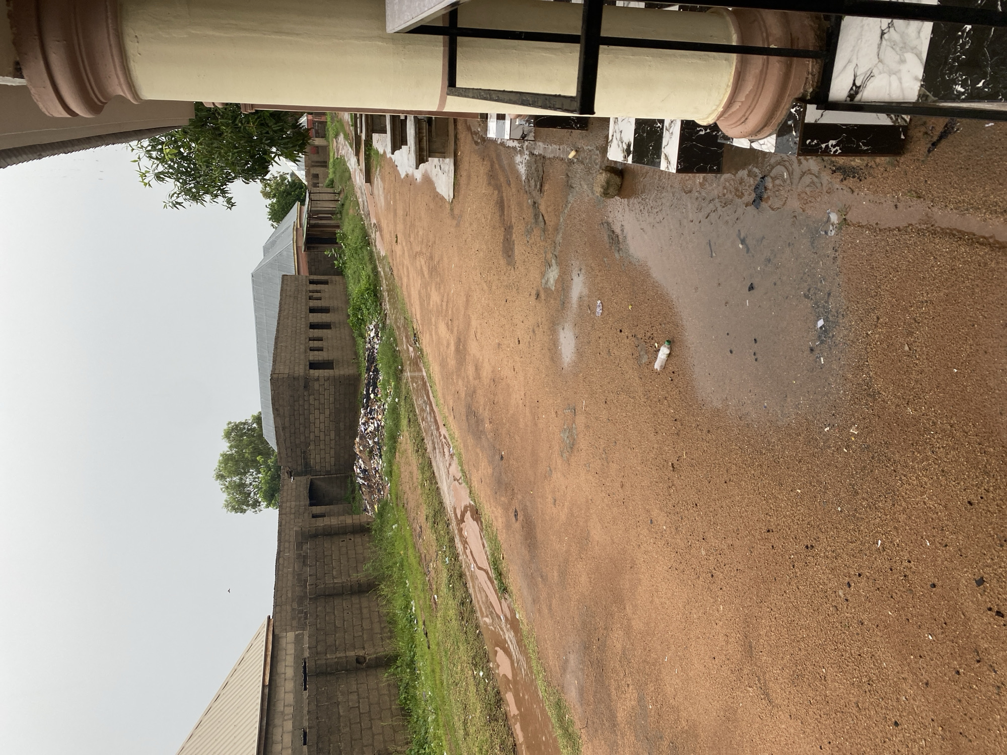 The image depicts a street scene in Jalingo, Nigeria, on a cloudy day, possibly during or after rainfall, as evidenced by puddles and water runoff. The foreground features a dirt road with patches of standing water and scattered debris, including plastic bottles. To the left, a low brick wall runs along the property, with overgrown grass and a pile of refuse behind it. Beyond the wall, several unfinished brick buildings with corrugated metal roofs are visible. To the right, a covered walkway or building exterior is present, supported by a large, cream-colored column with reddish-brown trim. The ground in this area is tiled with a black and white checkered pattern. The overall impression is one of a developing or perhaps neglected urban environment.