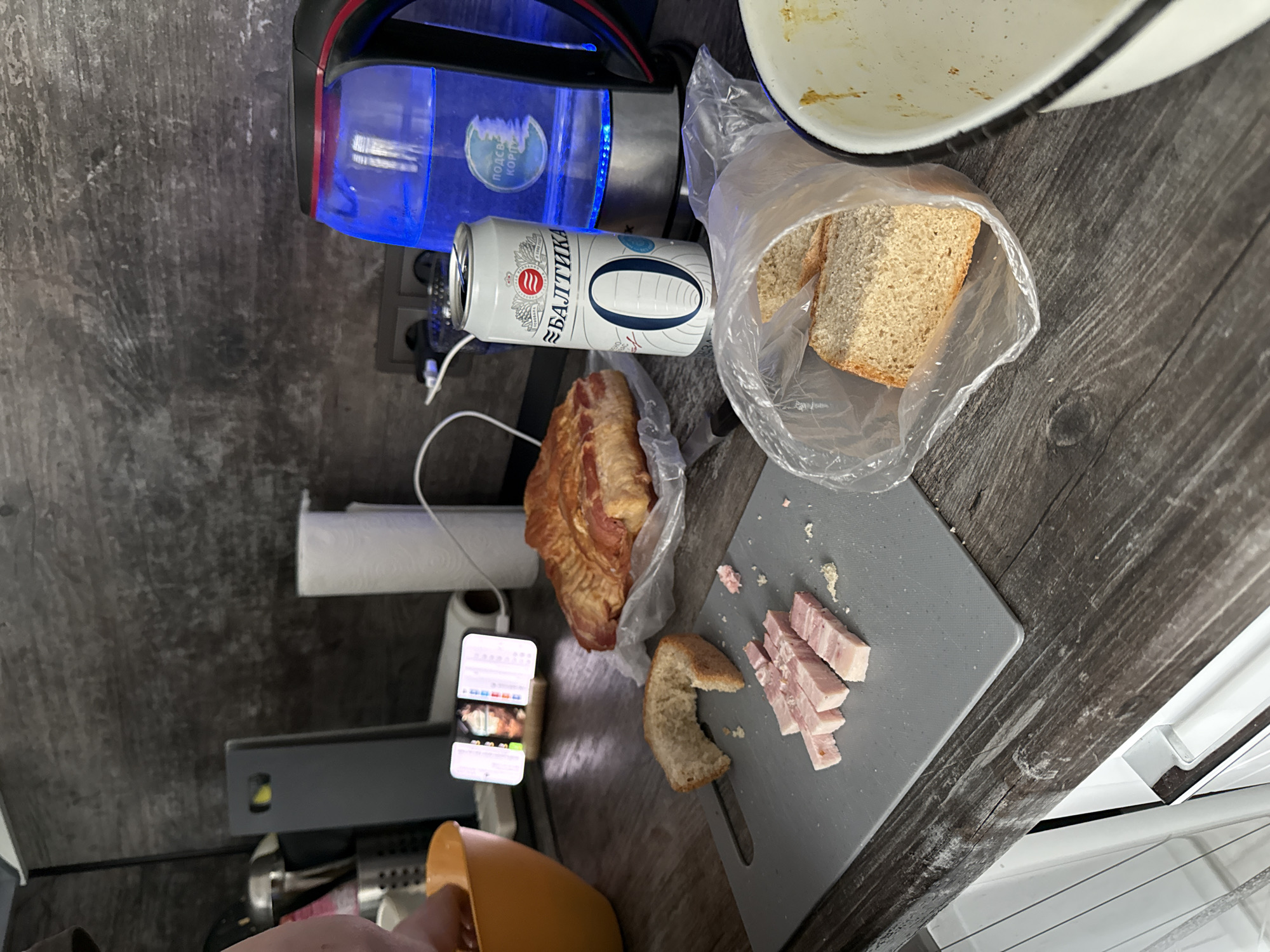 A cluttered countertop scene includes a blue electric kettle, a partially full can of Baltika 0 beer, and a plastic bag containing sliced bread. Nearby is a partially opened package of bacon and a gray cutting board with sliced bacon and a piece of bread. A smartphone on a stand is visible, and a person's hand is holding an orange object. The environment suggests an indoor kitchen or food preparation area. No specific location within Moscow is discernible from the image.