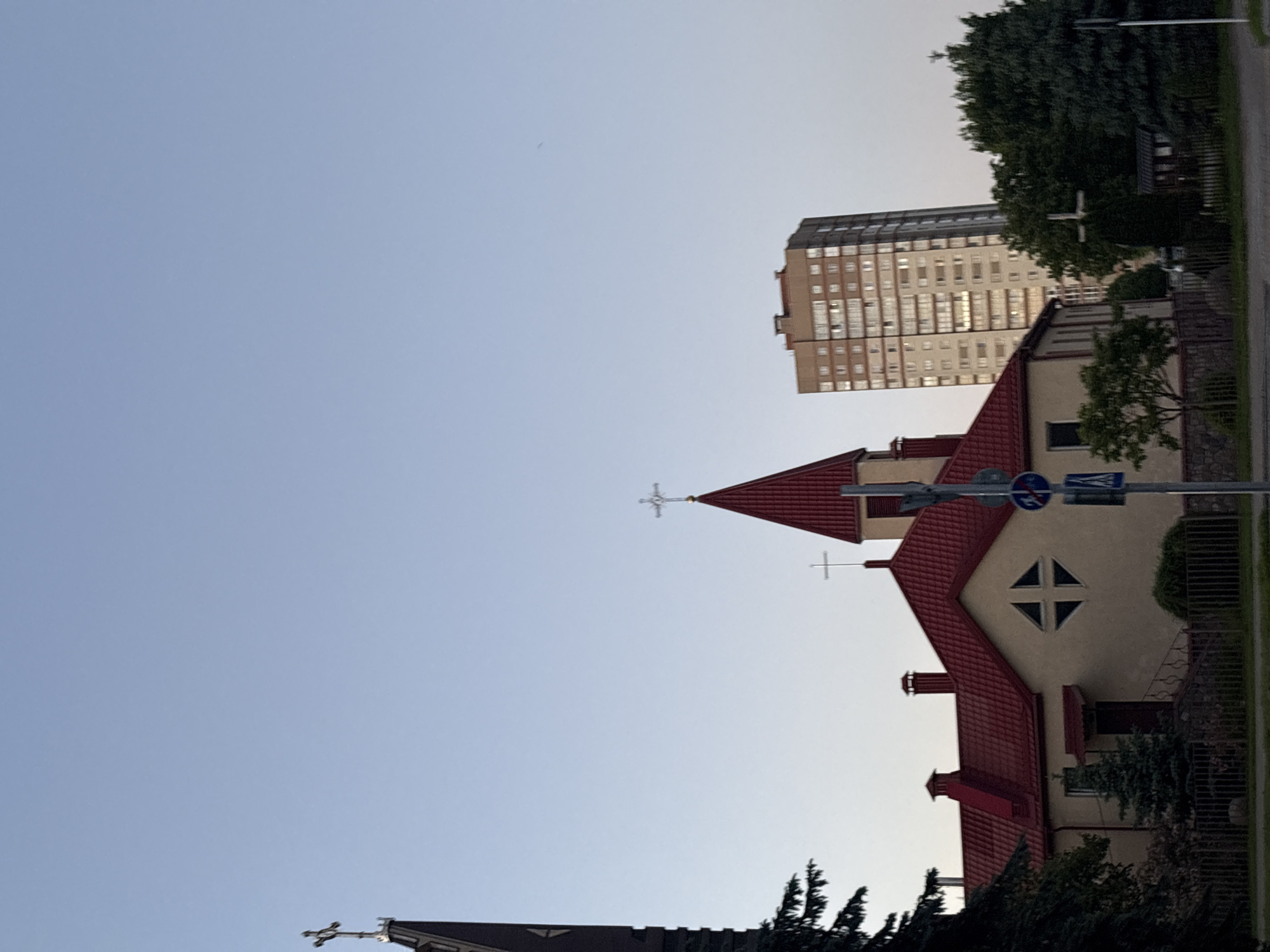 The image shows a church with a red roof and white walls in Minsk, Belarus, juxtaposed with a modern high-rise apartment building. The sky is clear, and the surrounding area includes trees and a street sign. The composition is vertically oriented.