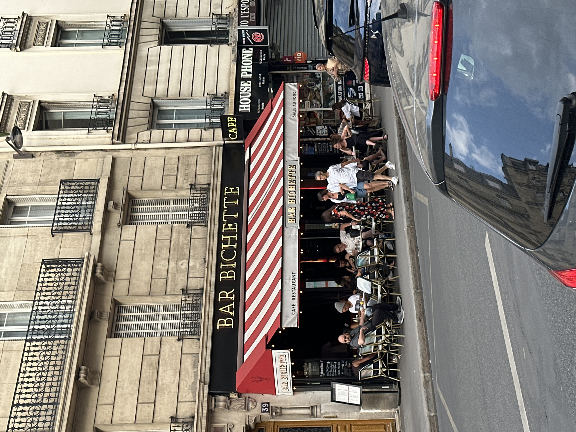 In Paris, France, an outdoor seating area of "Bar Biquette" cafe restaurant is visible. Red and white striped awnings cover the outdoor dining space where several patrons are seated at small tables. A black menu board is positioned to the left of the entrance. Adjacent to the cafe, a "House Phone" store is open for business. The street in front of the establishments features a sidewalk and a road with a white line marking. A black car is parked on the right side of the frame, with its rear visible. The architecture of the surrounding buildings is typical of Parisian style, with stone facades and ornate balconies.