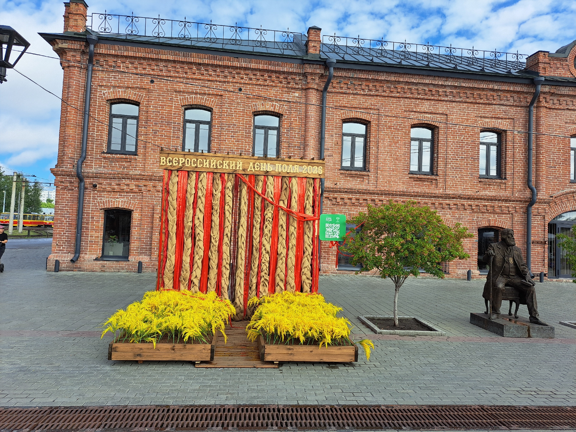 The image depicts a public square in Barnaul, Russia, outside a red brick building with arched windows. In the foreground, two wooden planters overflow with vibrant yellow foliage. A decorative backdrop, made of vertical straw-like elements interwoven with red fabric, stands behind the planters. A green sign with a QR code and Cyrillic text, reading "ДЕНЬ ПОЛЯ 2026" (Field Day 2026), is affixed to the backdrop. To the right, a bronze statue of a seated man with a beard and cane is positioned next to a small tree. The ground is paved with gray interlocking bricks. In the background, the red brick building, likely a historical structure, features a decorative wrought-iron railing along its roofline. The sky is partially cloudy. A person is visible on the left side of the image, riding a scooter.